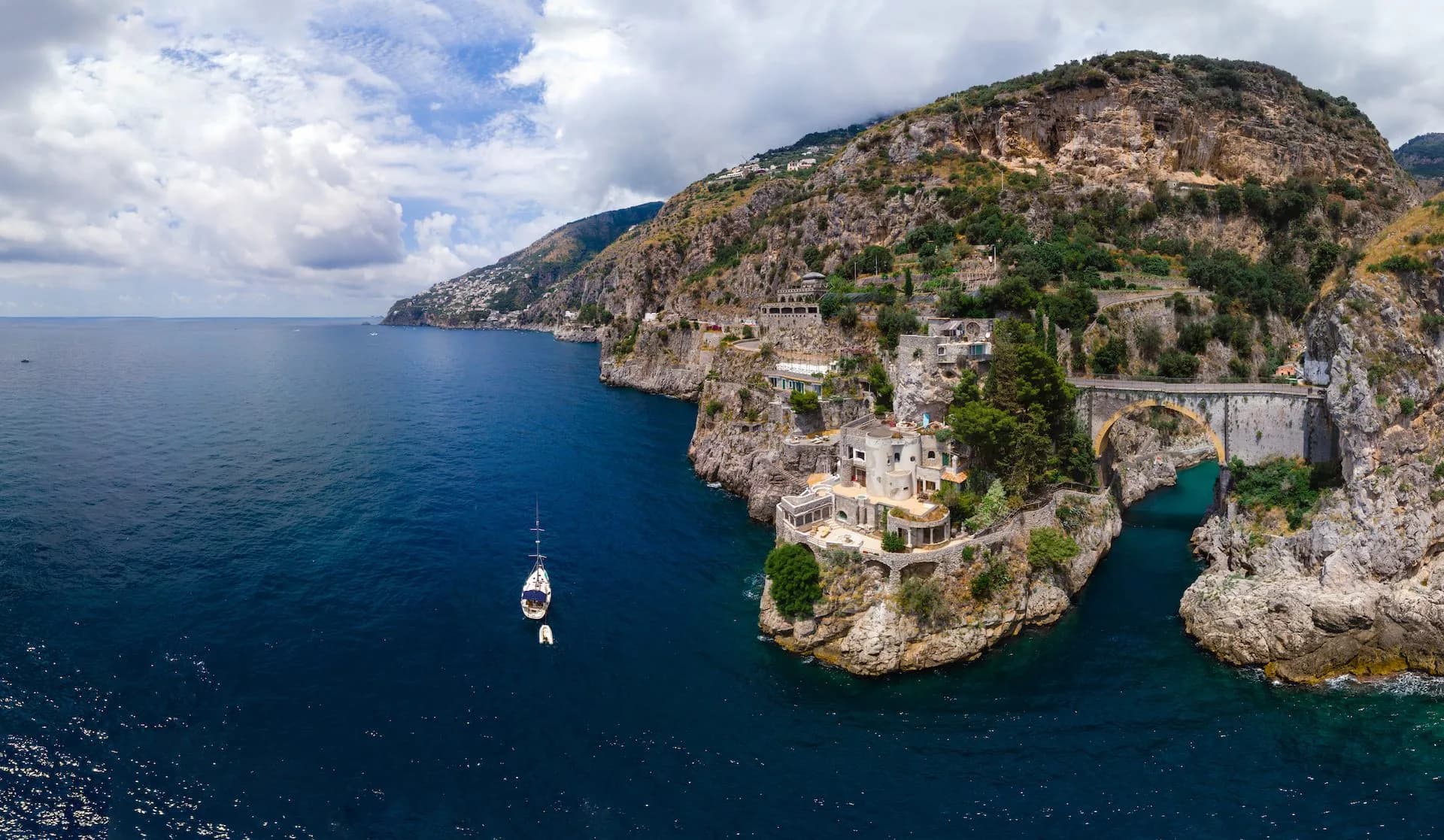 Sailboat on deep blue sea near Furore fjord with cliffside buildings and stone bridge.