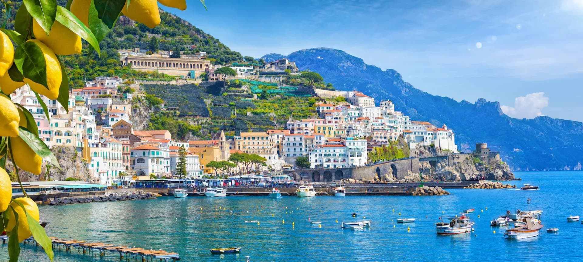 Lemons hang over Amalfi coast town built on steep green hillsides above blue water with boats.