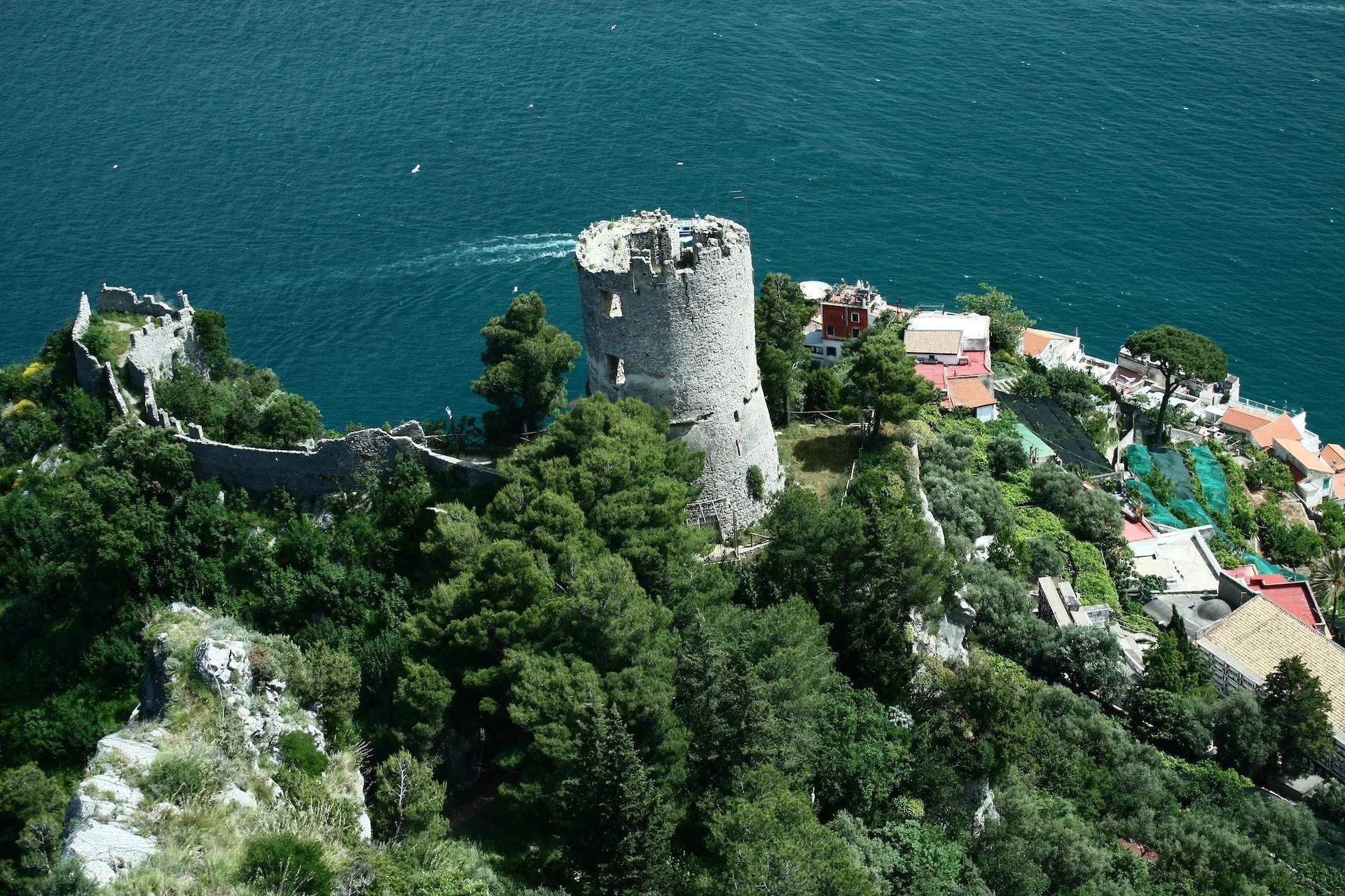 Ruined stone tower and walls on a lush green cliff overlooking deep blue sea near coastal village.