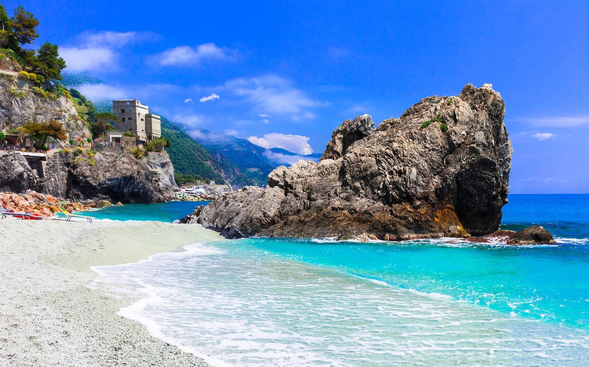 Gravel beach with turquoise water, large rock formation, and historic tower in Monterosso, Italy