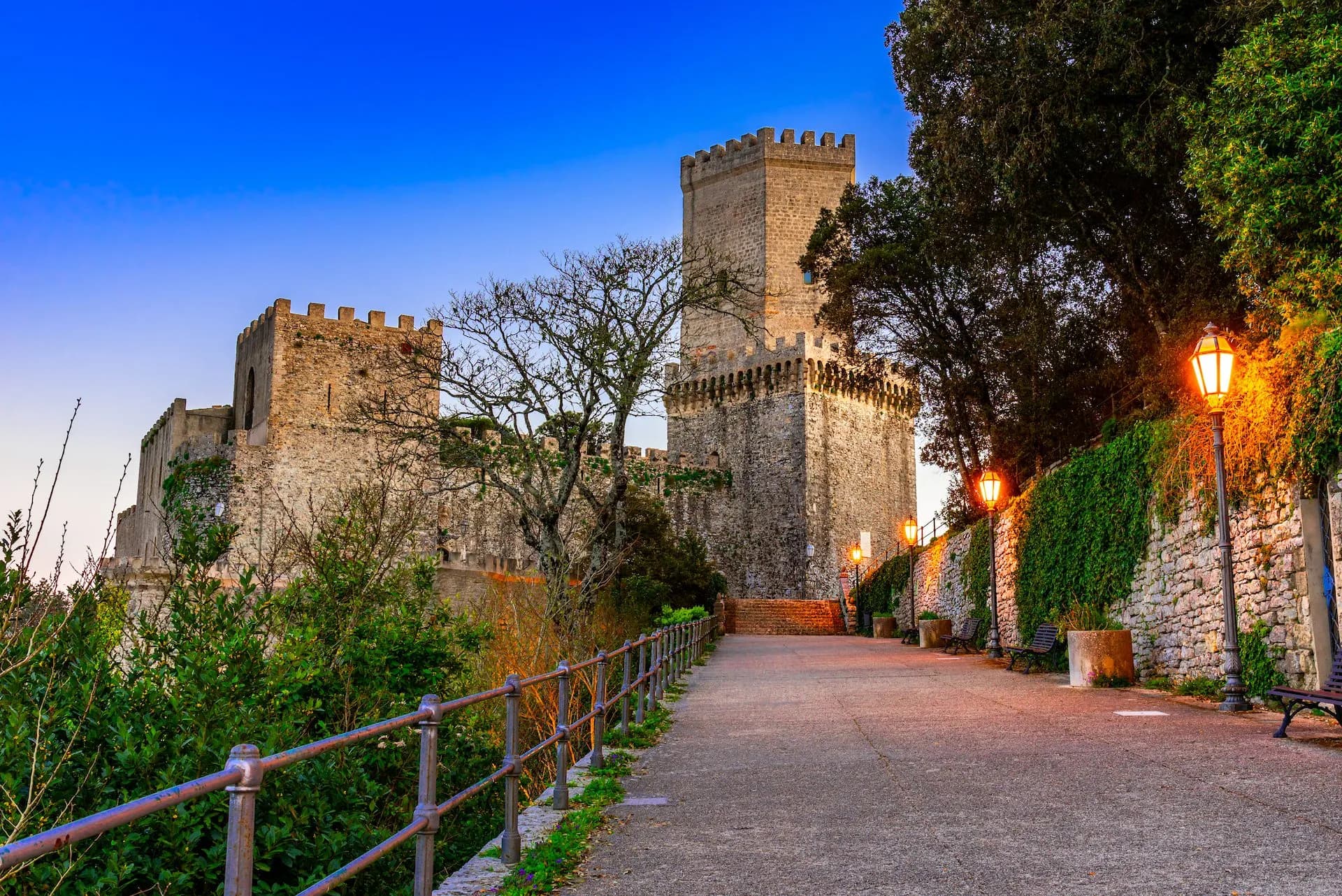 Stone castle walls and towers next to a path with illuminated lanterns at dusk in Erice.