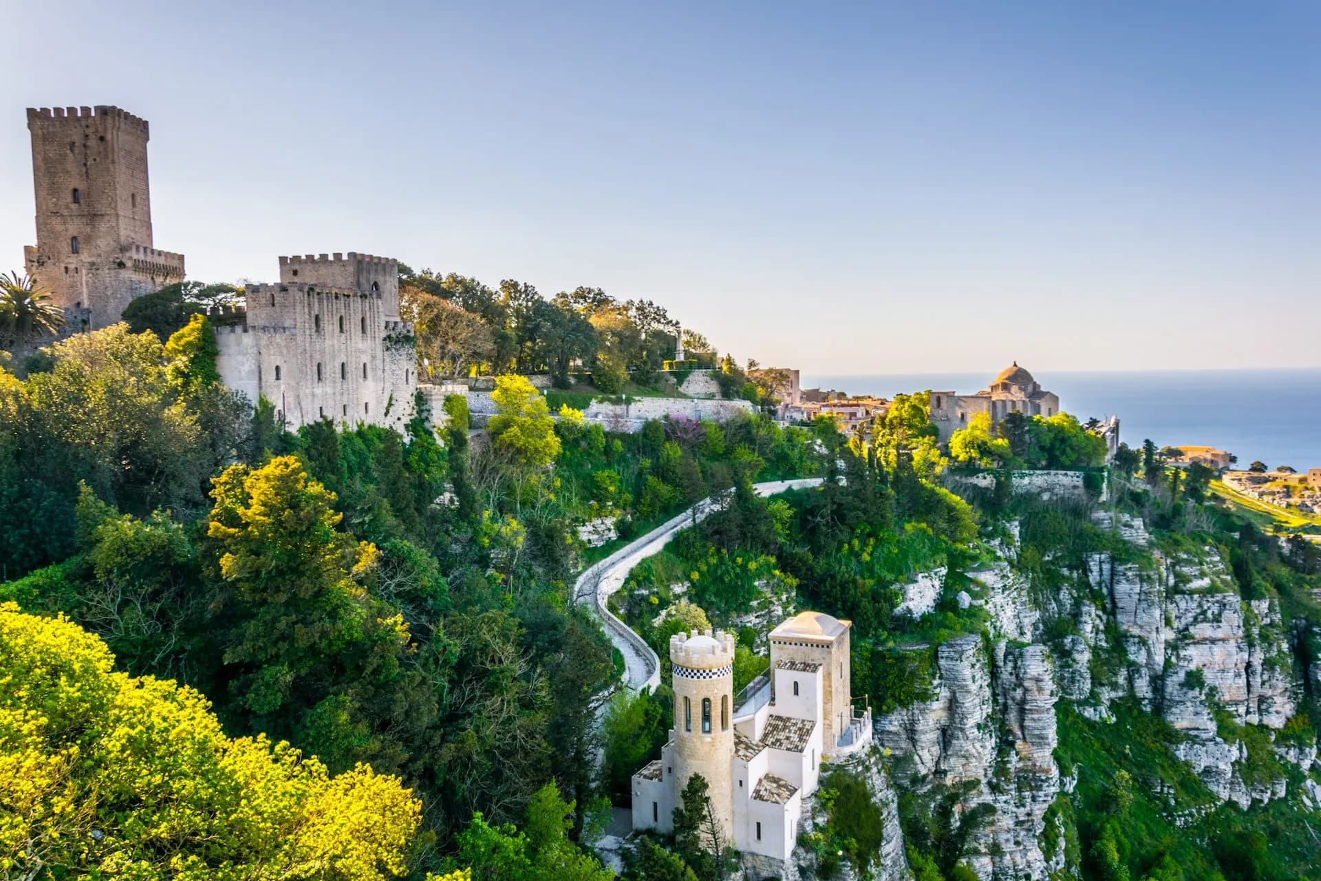Historic stone castles atop a lush, green cliff overlooking the Mediterranean Sea in Erice, Sicily.