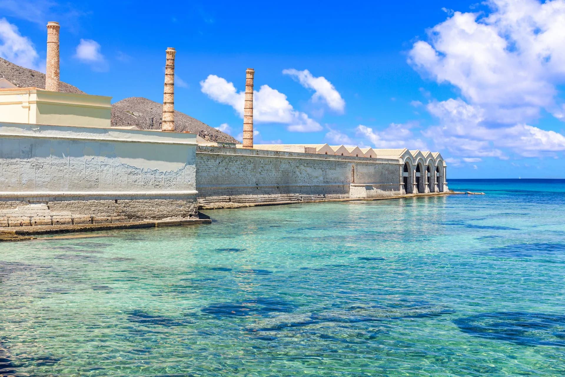Old tuna factory with brick chimneys on Favignana island by clear turquoise sea.