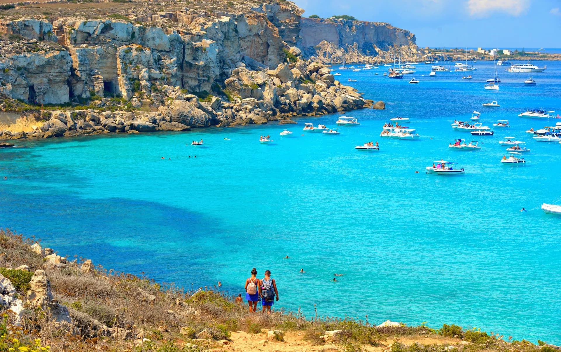 Boats anchored in turquoise cove beside rocky cliffs, Cala Rossa, Sicily.