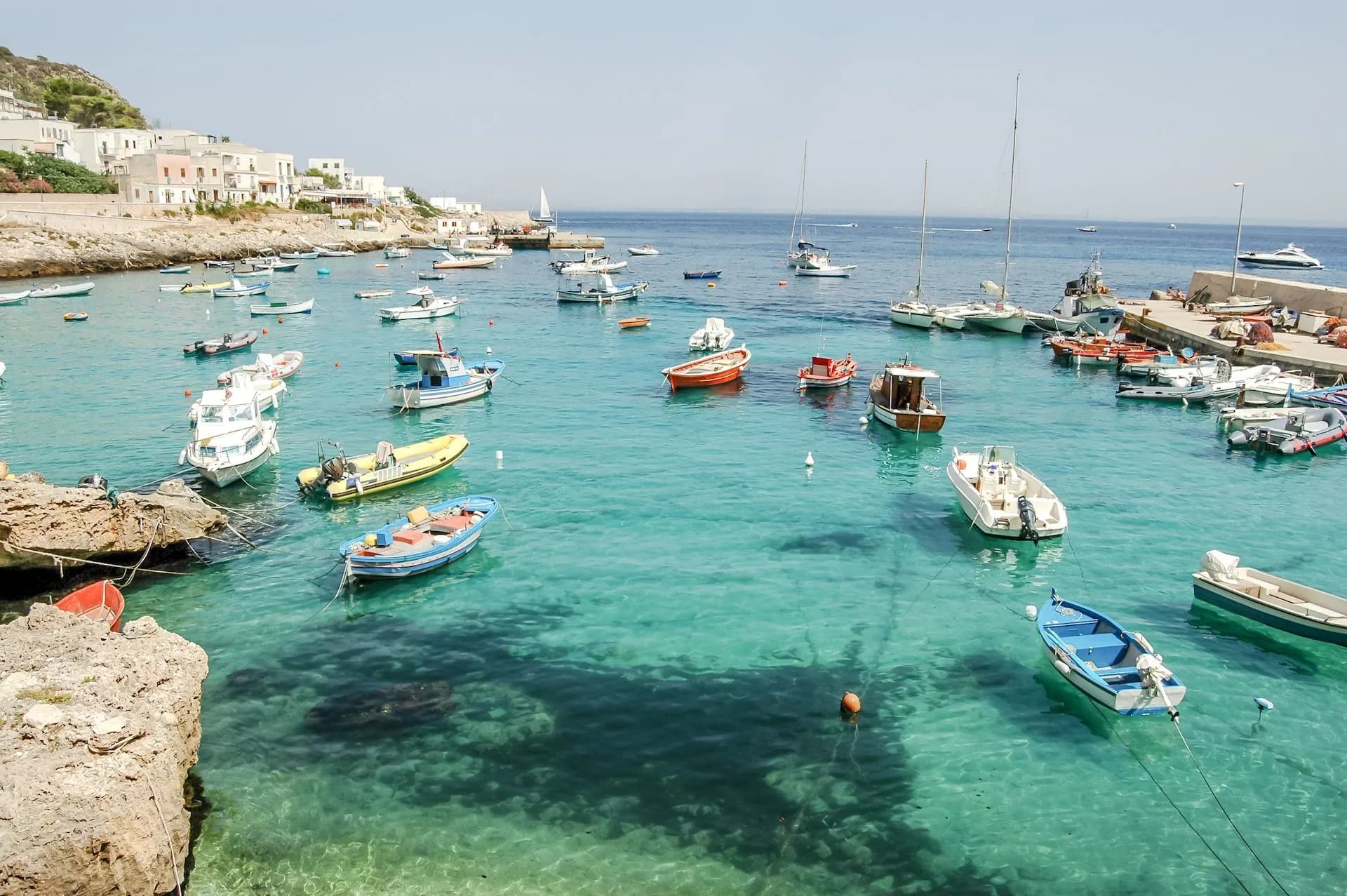 Numerous small boats anchored in turquoise harbor waters near white coastal buildings in Levanzo.