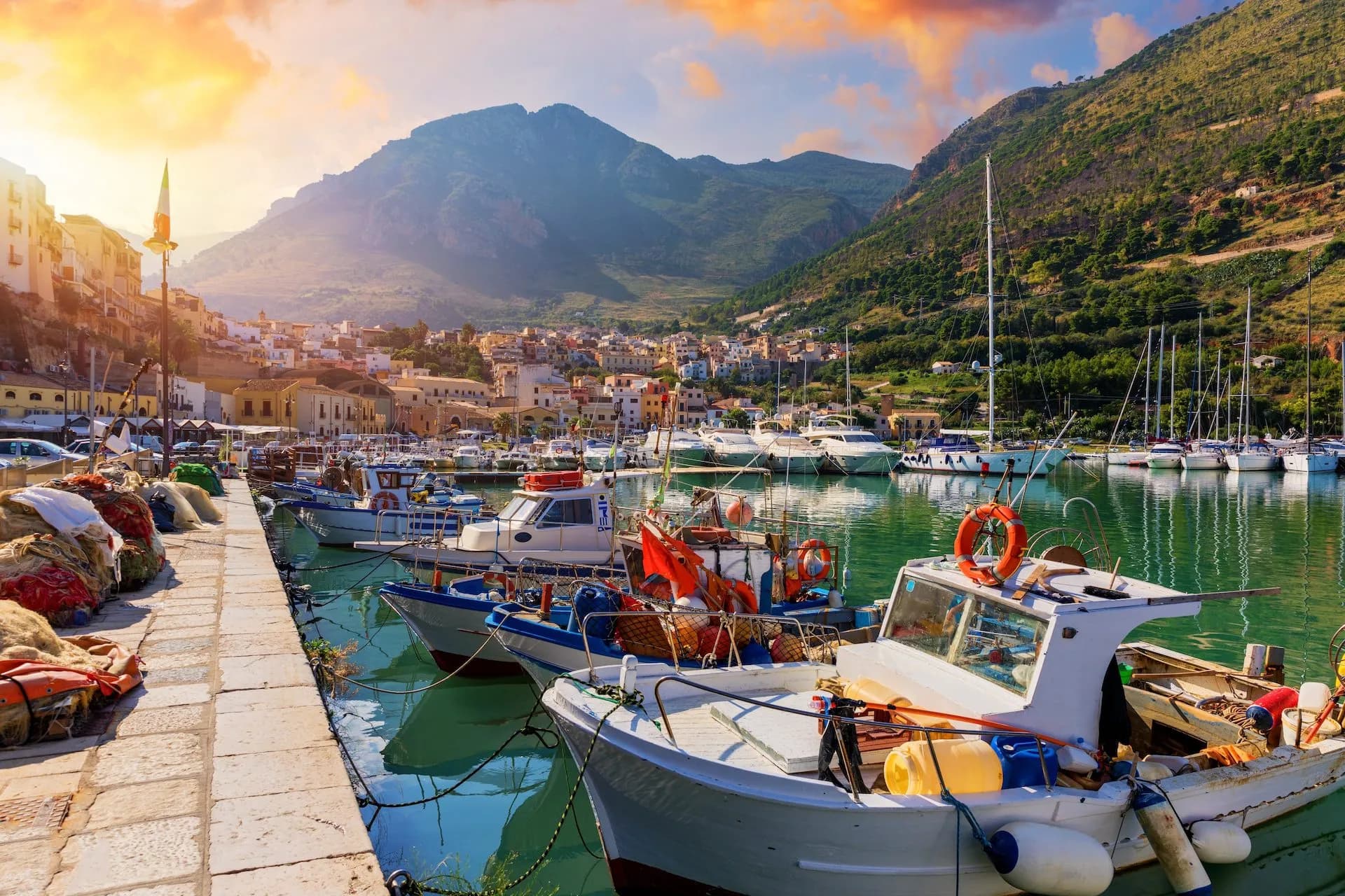 Fishing boats docked in Trapani harbor with colorful town and mountains at sunset
