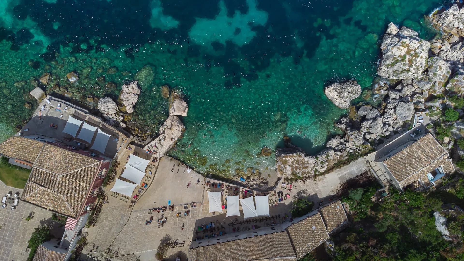 Aerial view of buildings and sunbathers near turquoise Mediterranean sea with rocky coastline in Sicily.