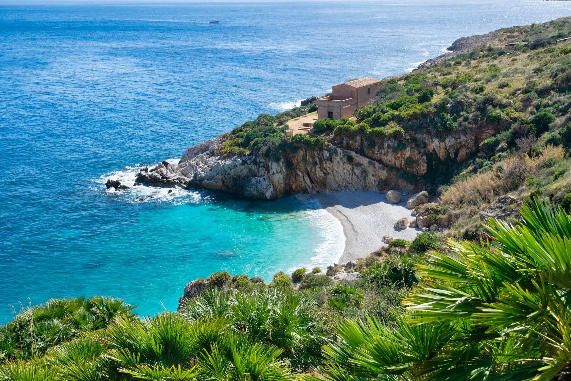 Secluded cove with turquoise water, small beach, and house on a rocky, green cliff in Zingaro Nature Reserve.