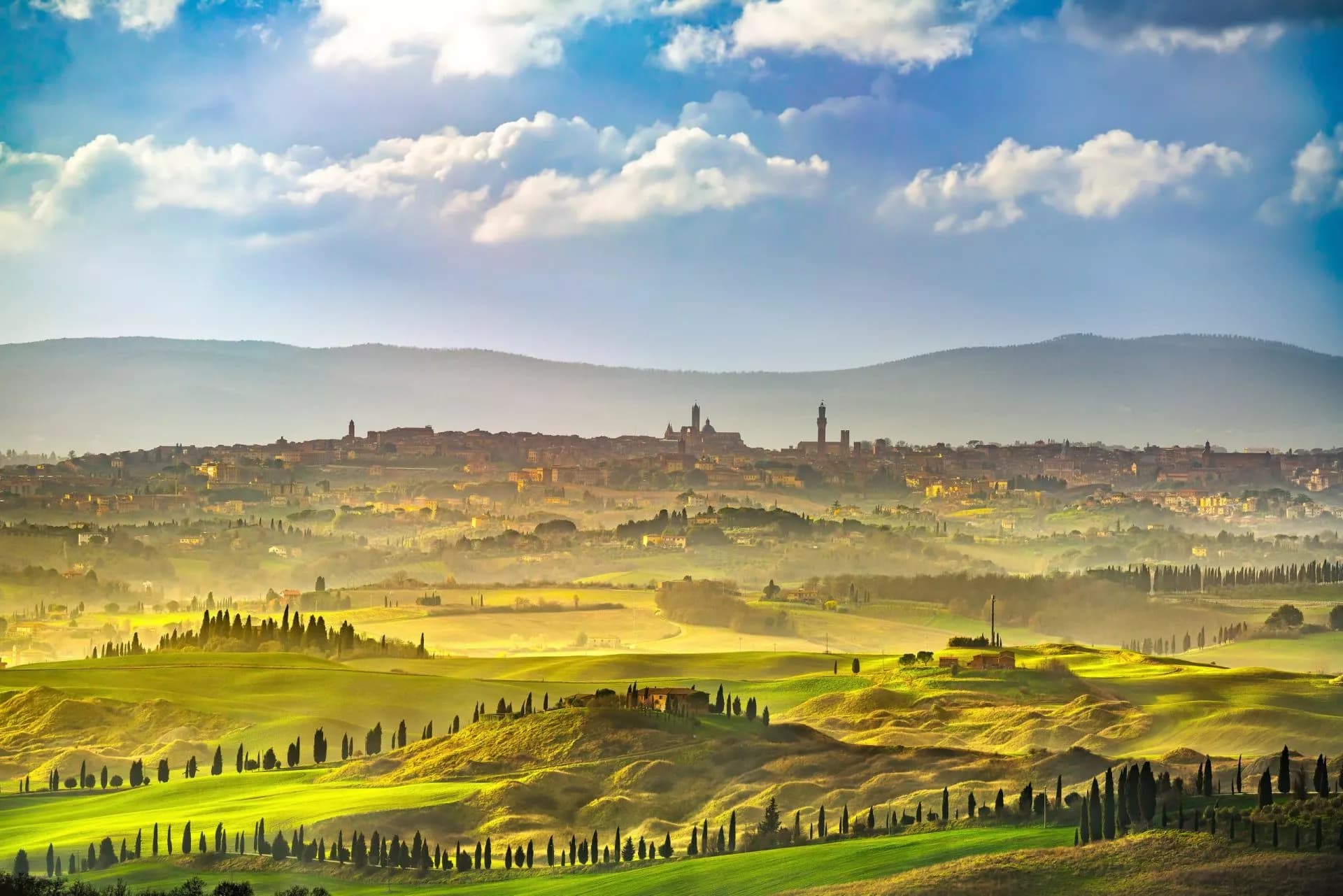 Siena city skyline above rolling green Tuscan hills with cypress trees under a cloudy blue sky.