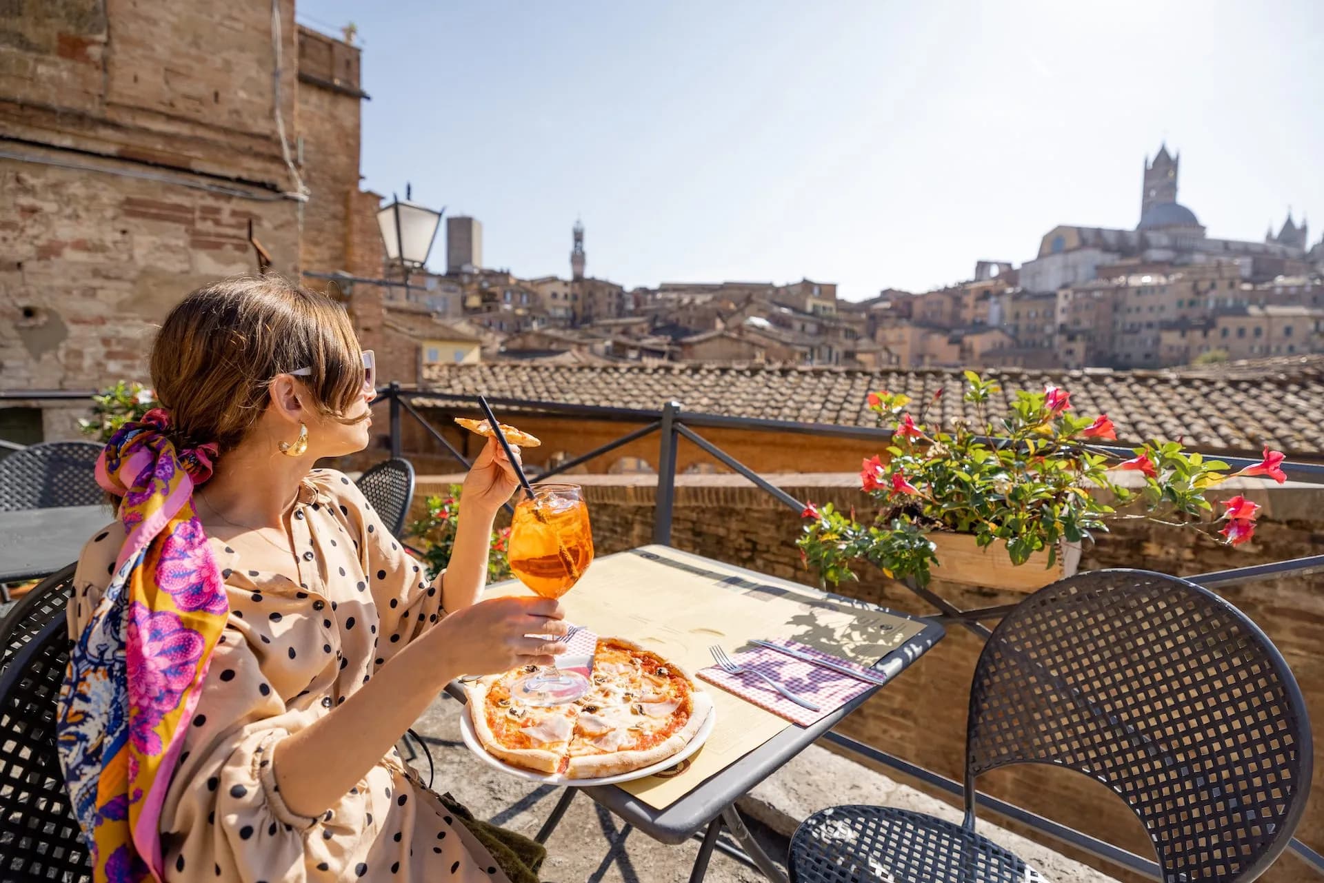 Woman dining on pizza and Aperol spritz on terrace overlooking historic Siena cityscape.
