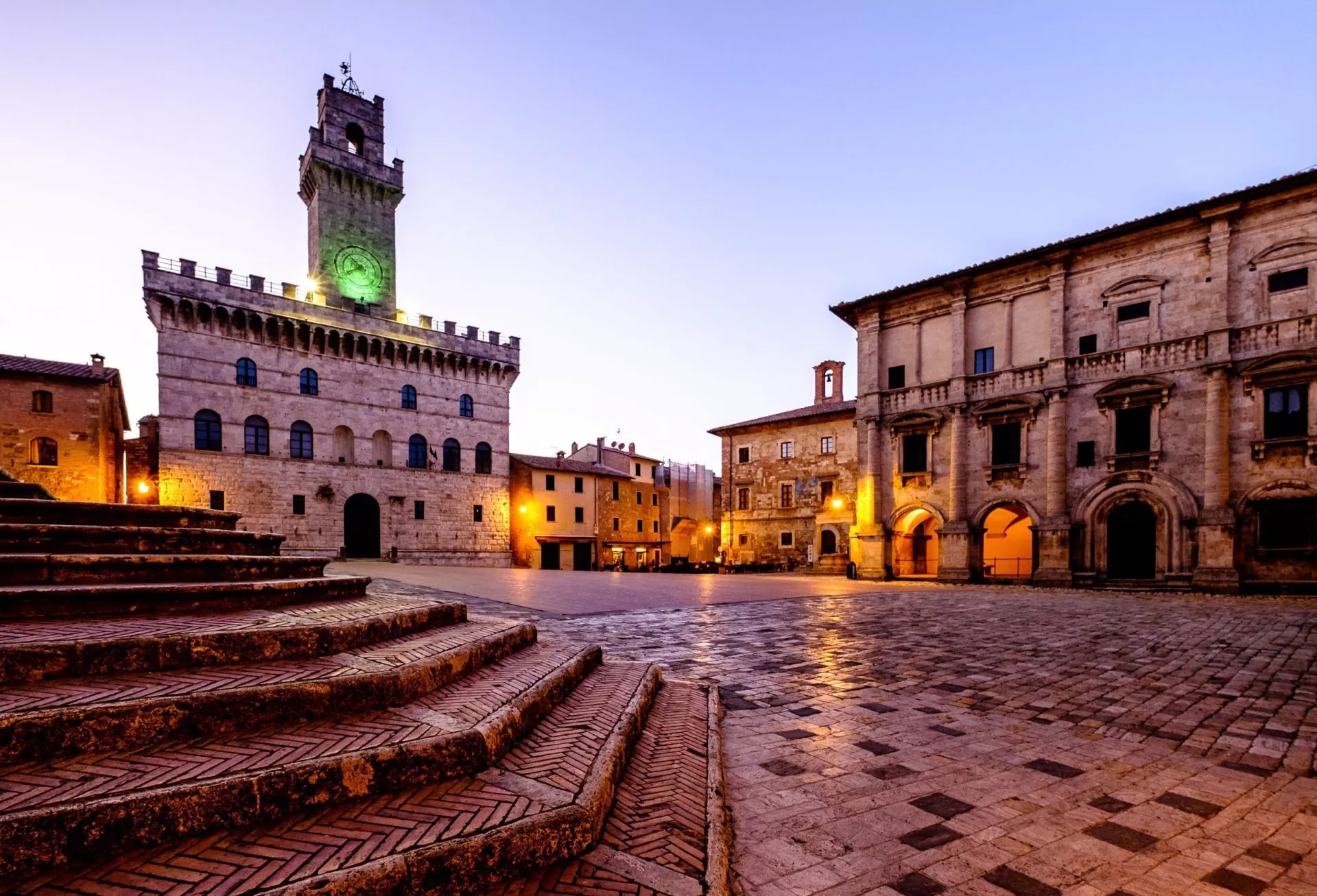 Piazza illuminated at dusk with historic stone buildings and clock tower in Montepulciano.