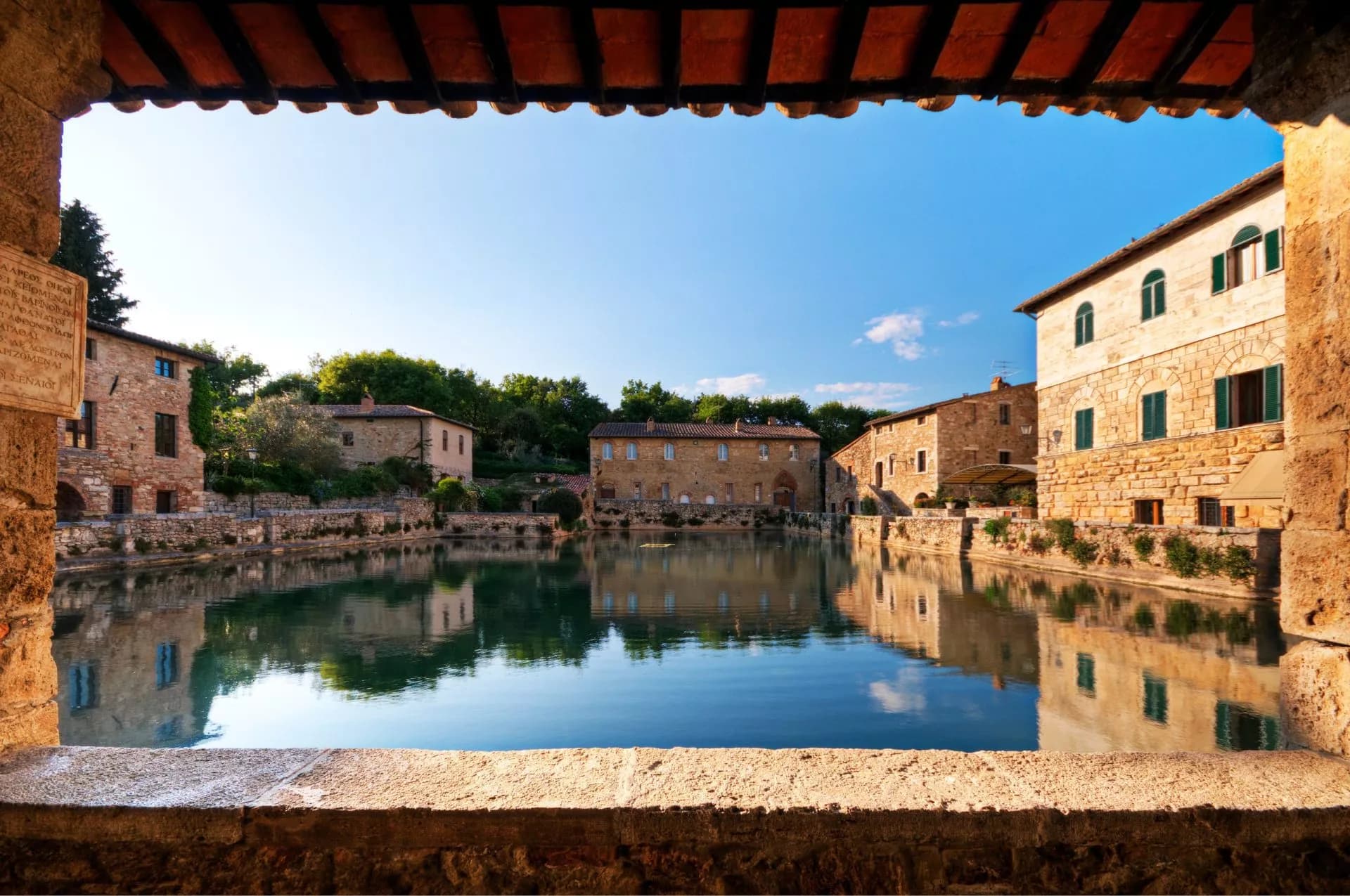 Thermal pool reflecting historic stone buildings, viewed through a stone archway in Bagno Vignoni.