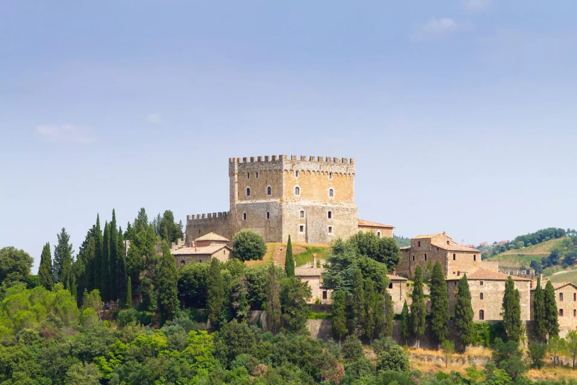 Ripa d'Orcia Castle fortress on a hill surrounded by cypress trees and lush greenery under a clear sky.