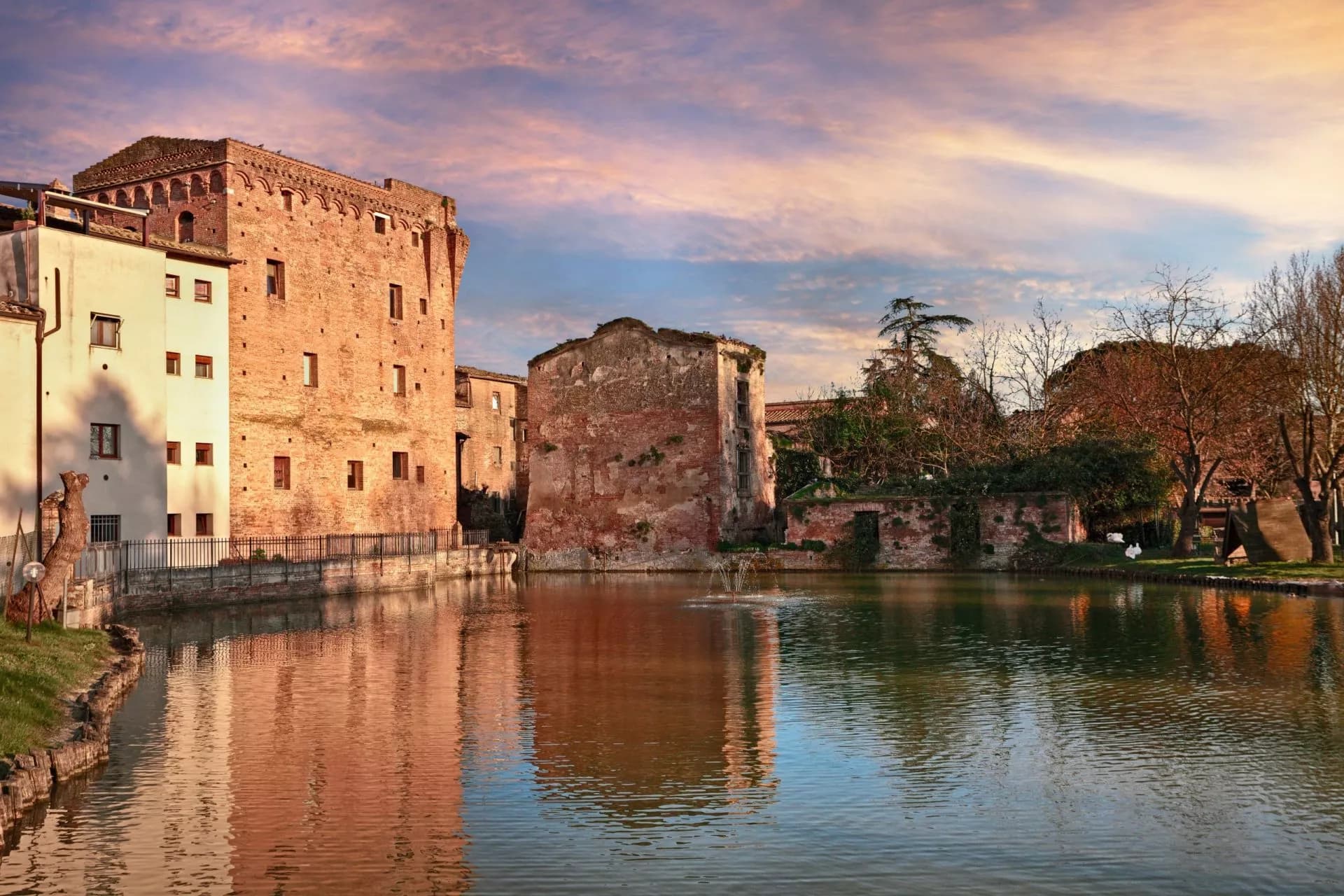 Historic brick buildings reflected in water with a small fountain under a colorful sunset sky.