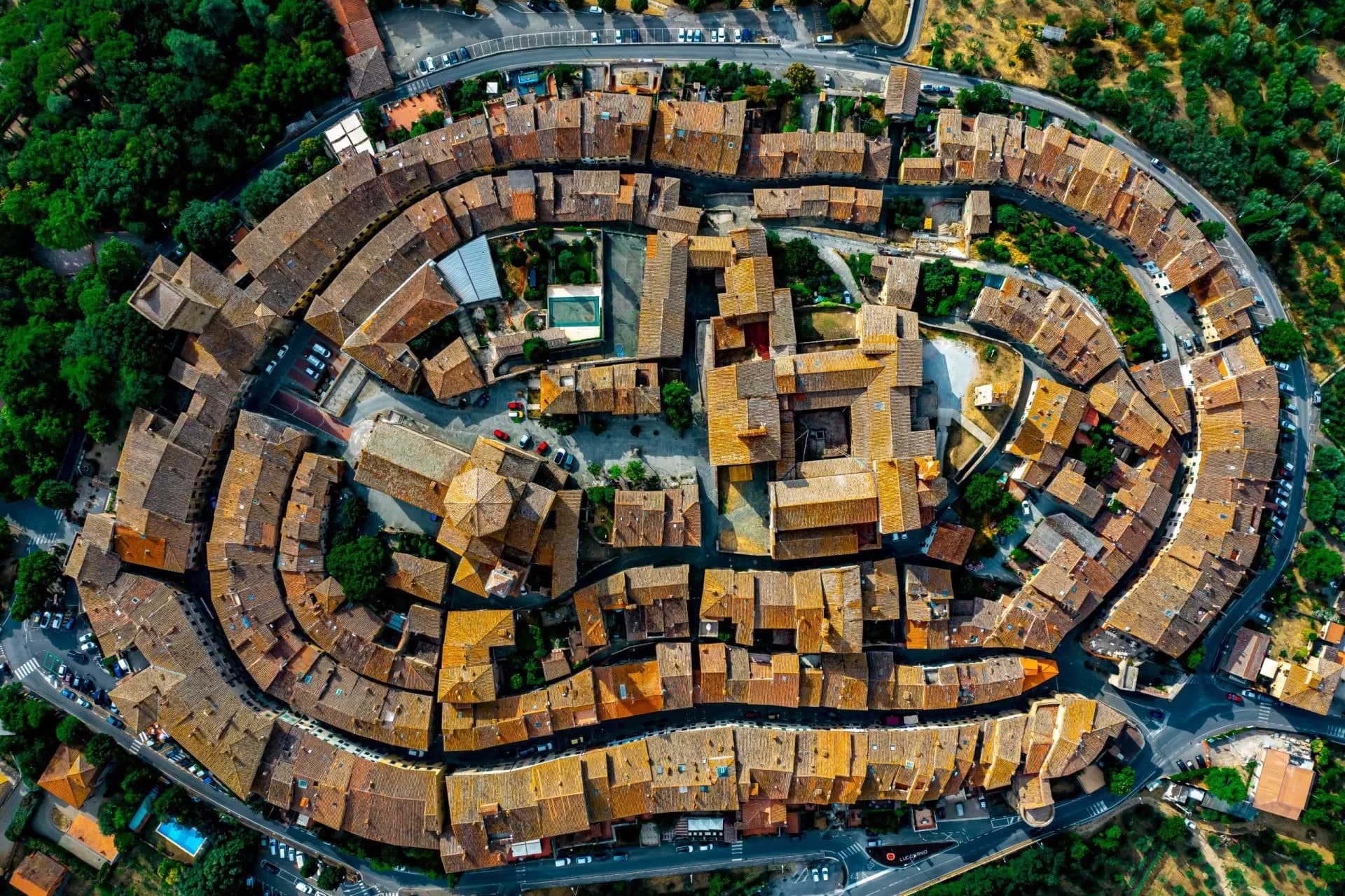Aerial view of the circular medieval town walls and terracotta roofs of Lucignano surrounded by green landscape.