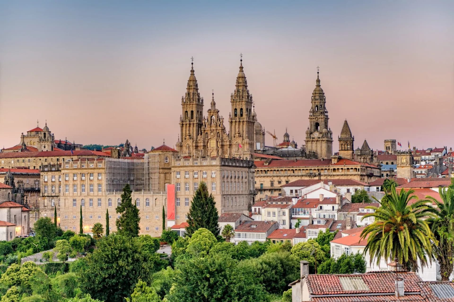 Hazy sunset over the monumental Santiago de Compostela Cathedral and city skyline with lush greenery.