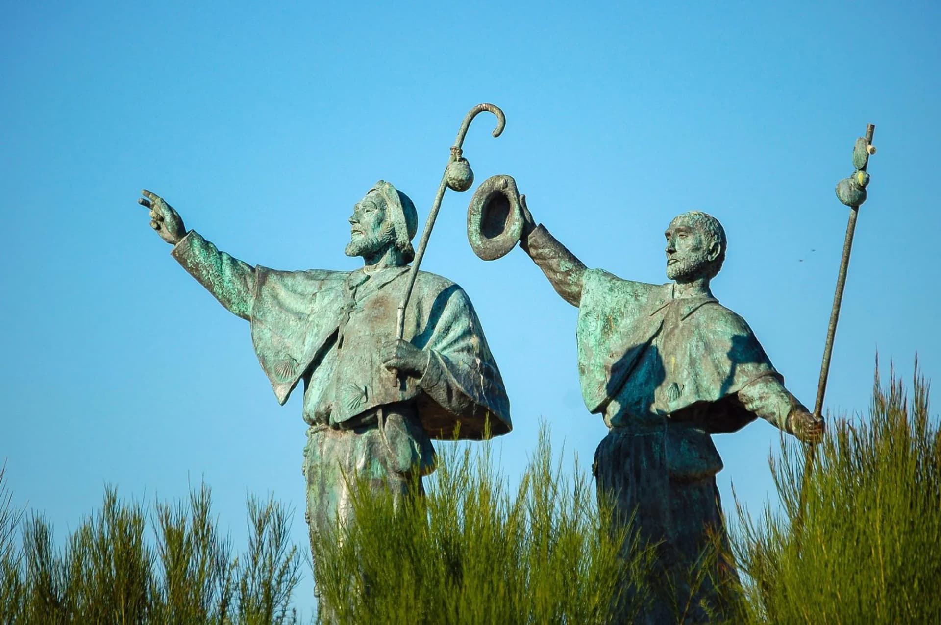 Bronze statues of pilgrims with staffs and hat against clear blue sky outside Santiago de Compostela.