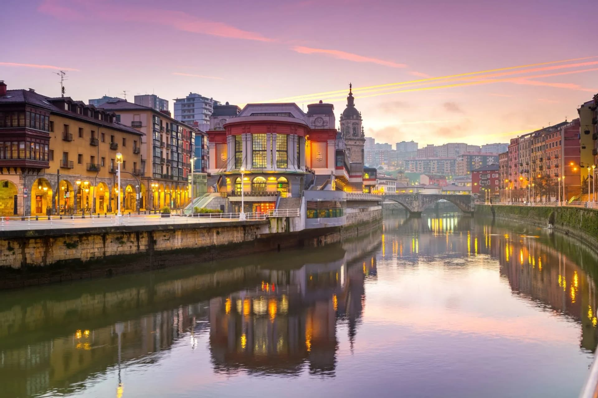 Ribera Market and illuminated buildings reflected in the river at dawn in Bilbao.