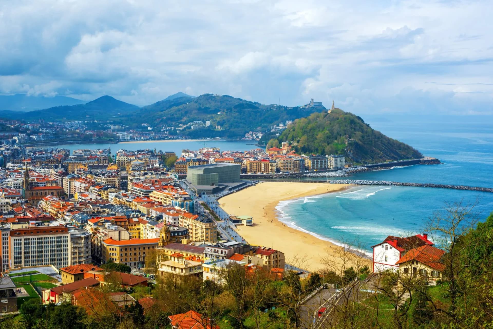 Panoramic view of San Sebastian city, La Concha beach, and green mountains under a cloudy sky.
