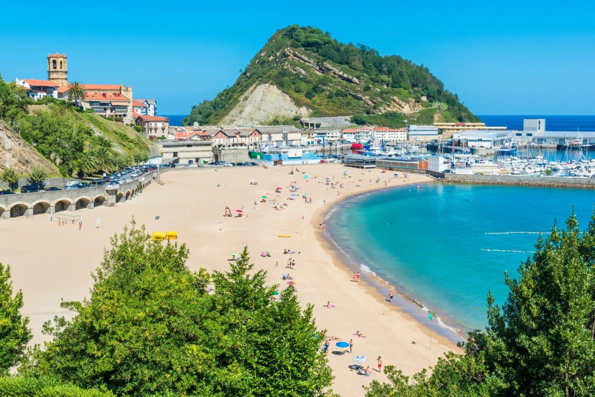Beachgoers on sand next to turquoise water, marina, and green hill in Getaria.
