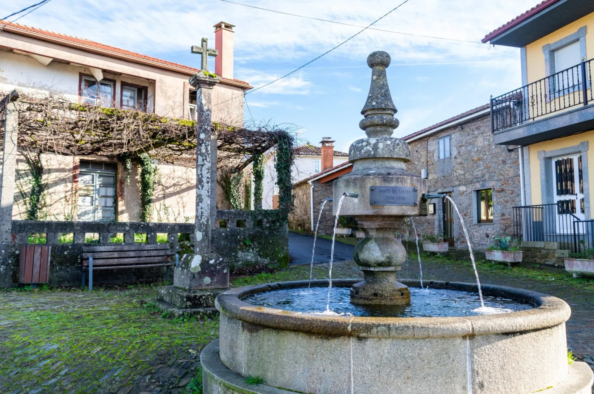 Stone water fountain in Melide square with mossy cobblestones and traditional houses