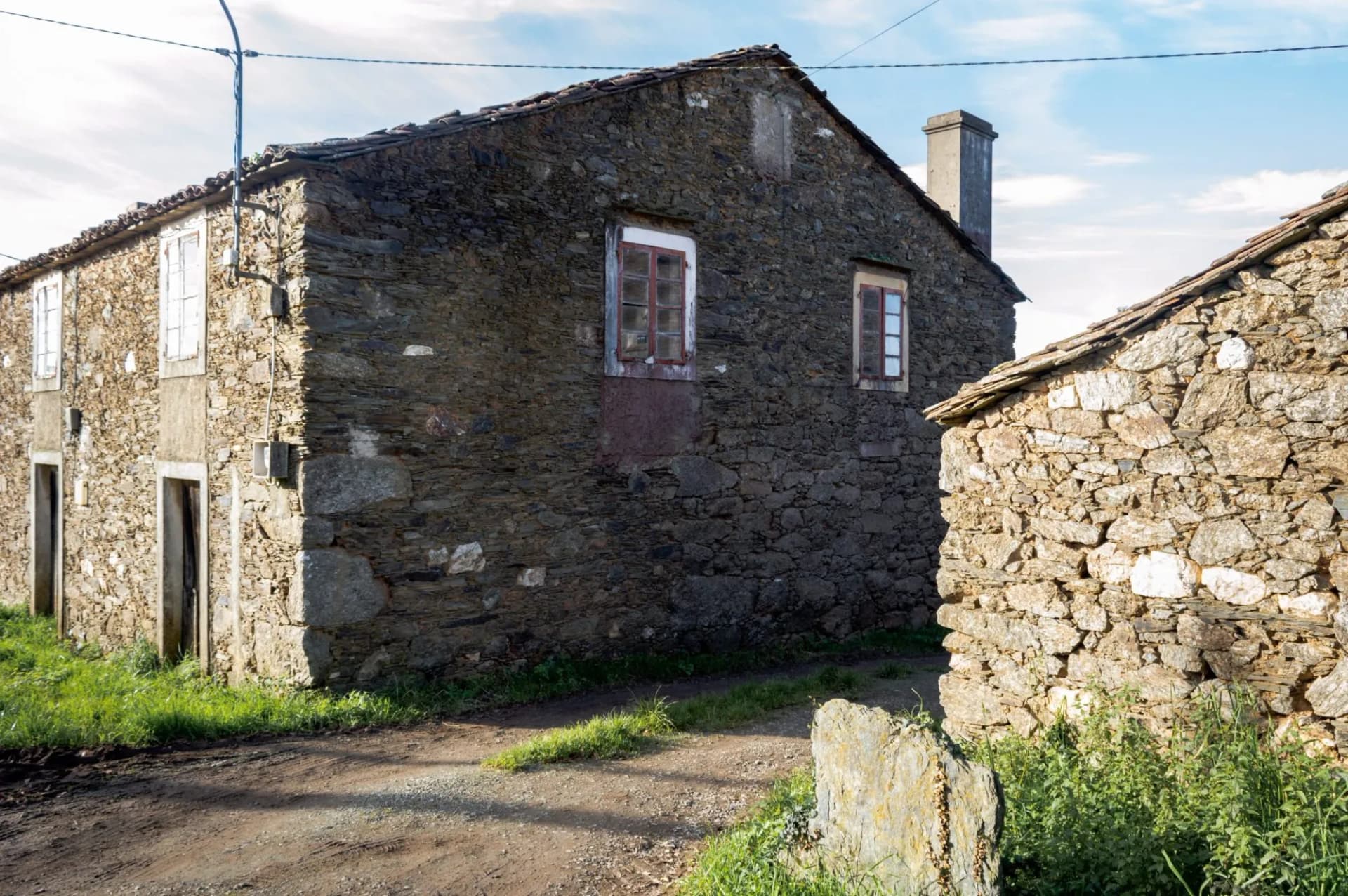 Stone houses with slate roofs and dirt path in Arzúa under a partly cloudy sky.