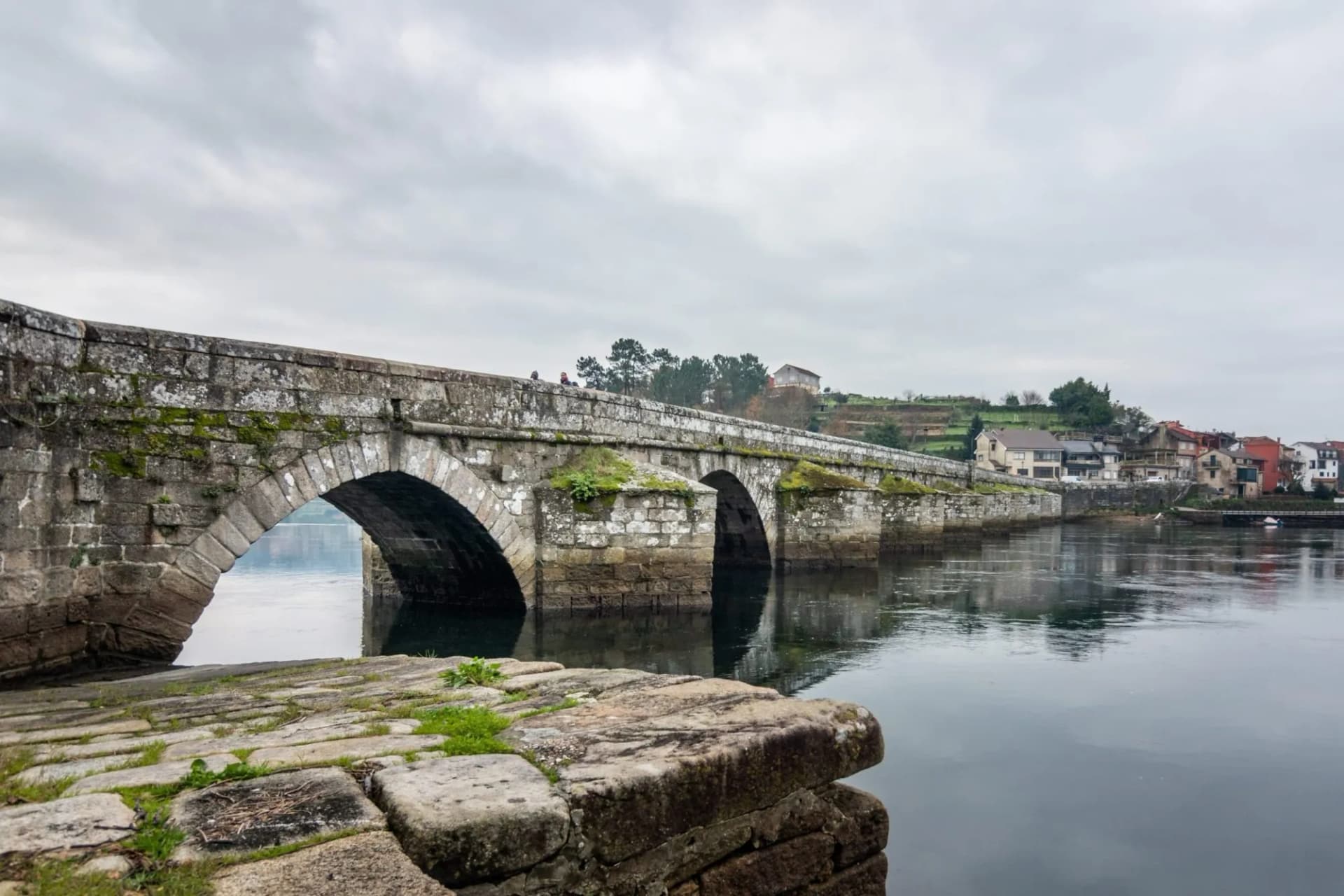 Stone bridge with mossy arches over dark water near Ponte Sampaio village under cloudy sky.