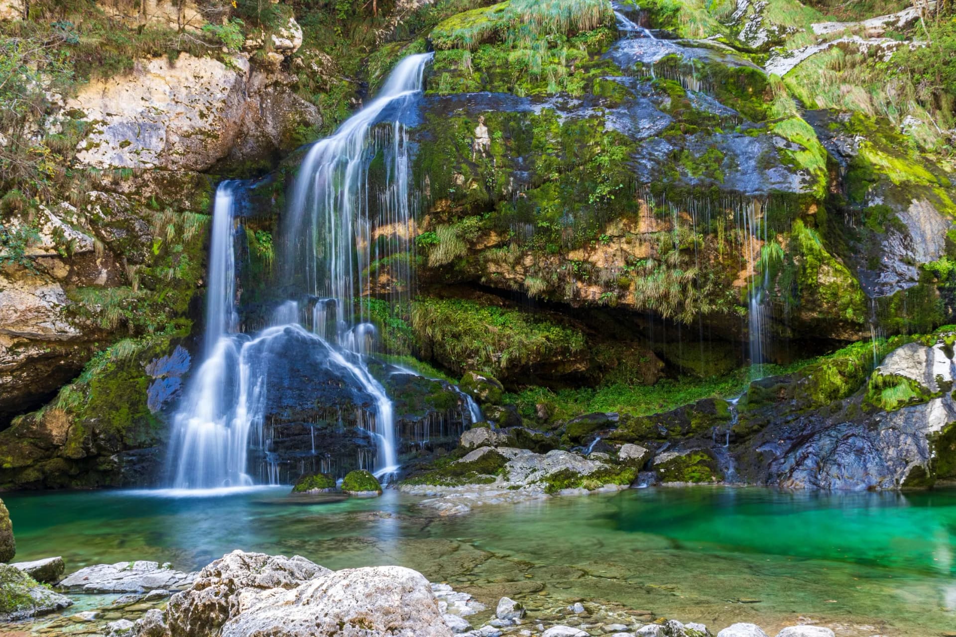 Virje Waterfall cascading over mossy rocks into a clear, emerald pool in a forest setting.