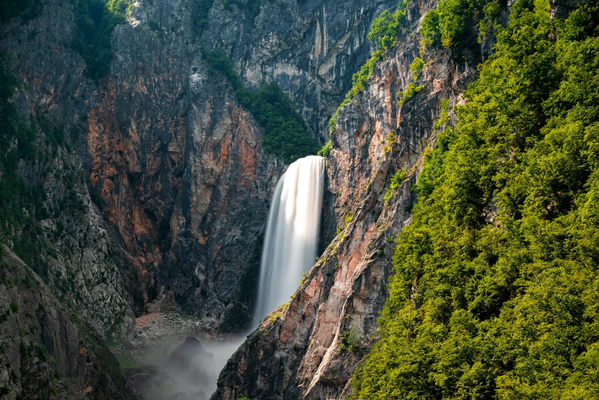 Tall waterfall plunging between steep, rocky cliffs covered in green trees, Boka Waterfall.