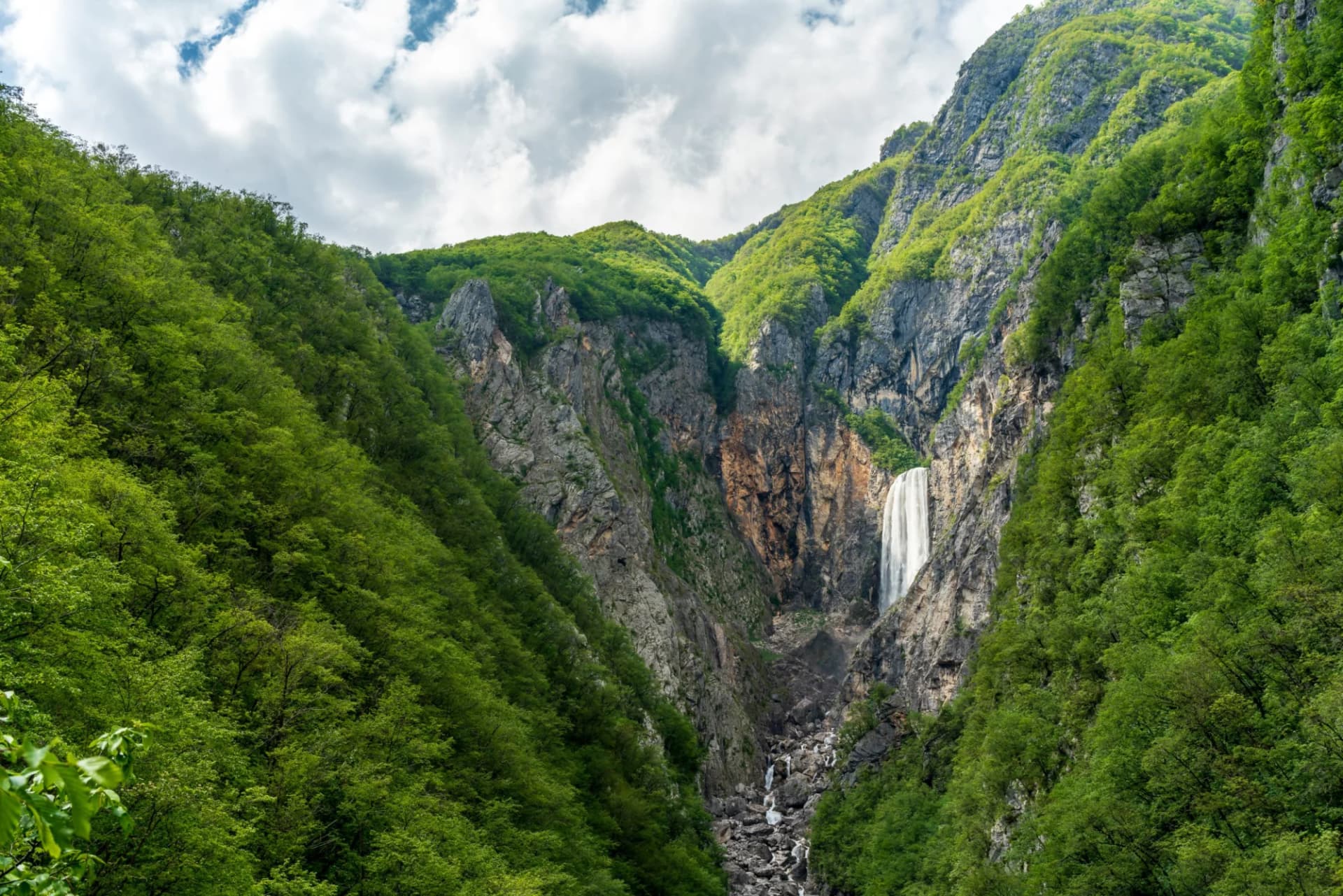 Tall waterfall cascading down sheer rock cliffs surrounded by lush green forested mountains in Slovenia.