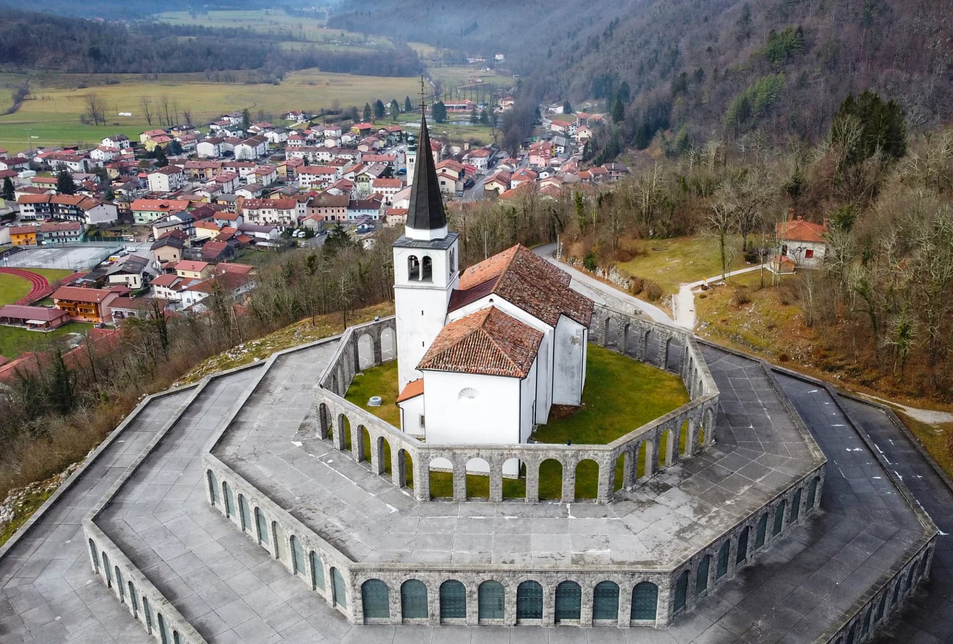 Aerial view of Italian Military Memorial church with white walls and stone arches overlooking a valley town.