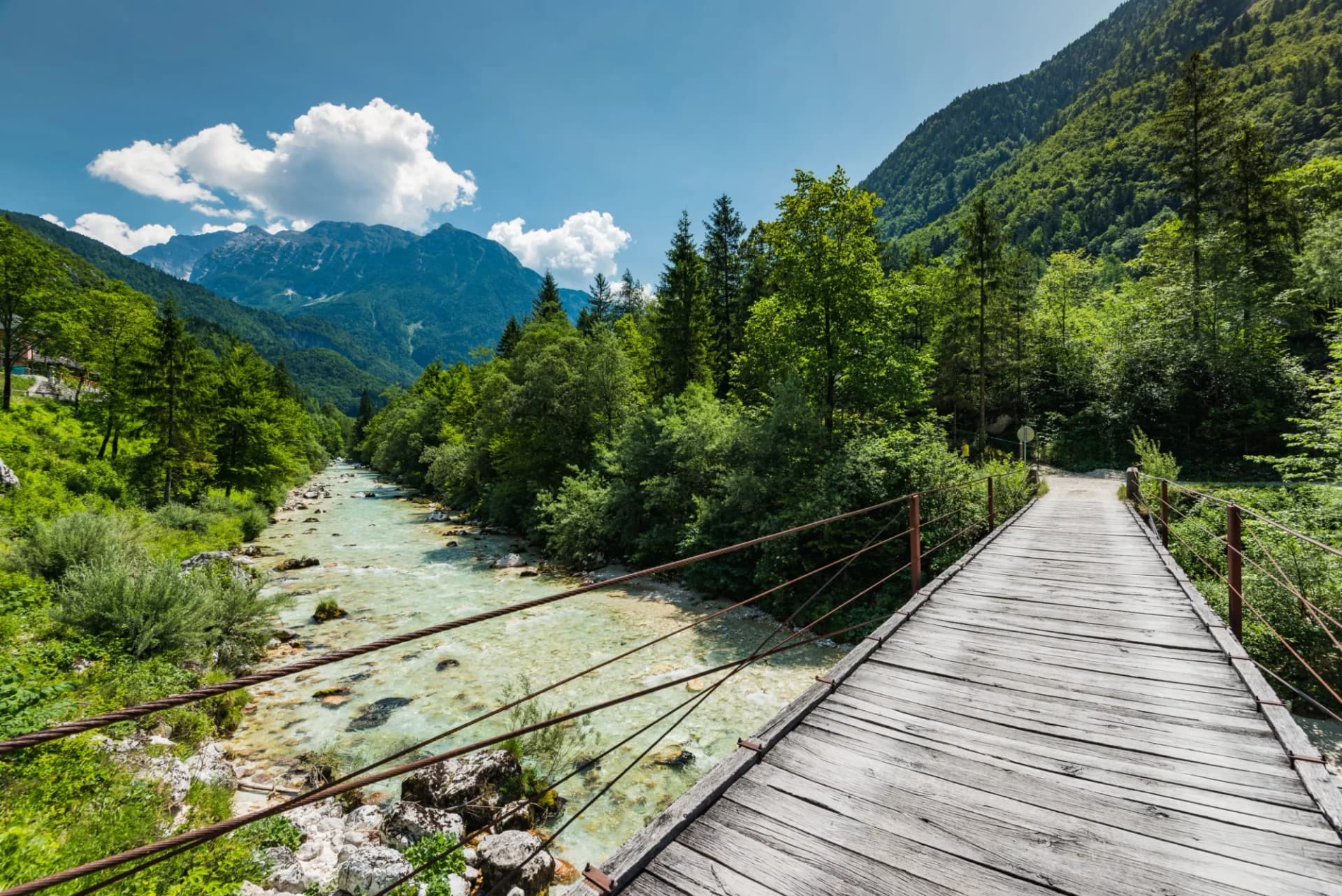 Wooden suspension bridge over the turquoise Soca River with forested mountains under a blue sky in Slovenia.