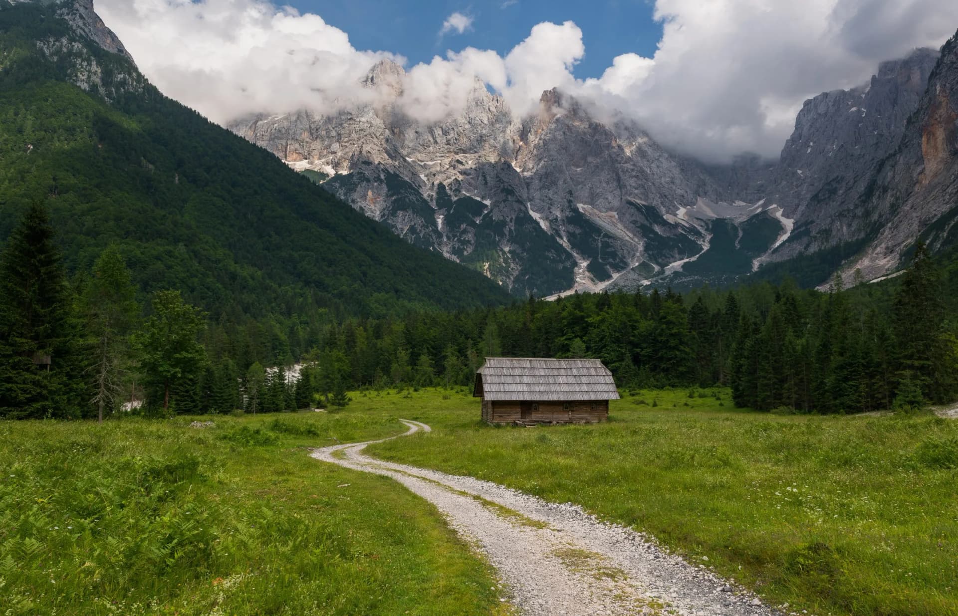 Wooden cottage in green meadow with gravel path, Julian Alps mountains in background