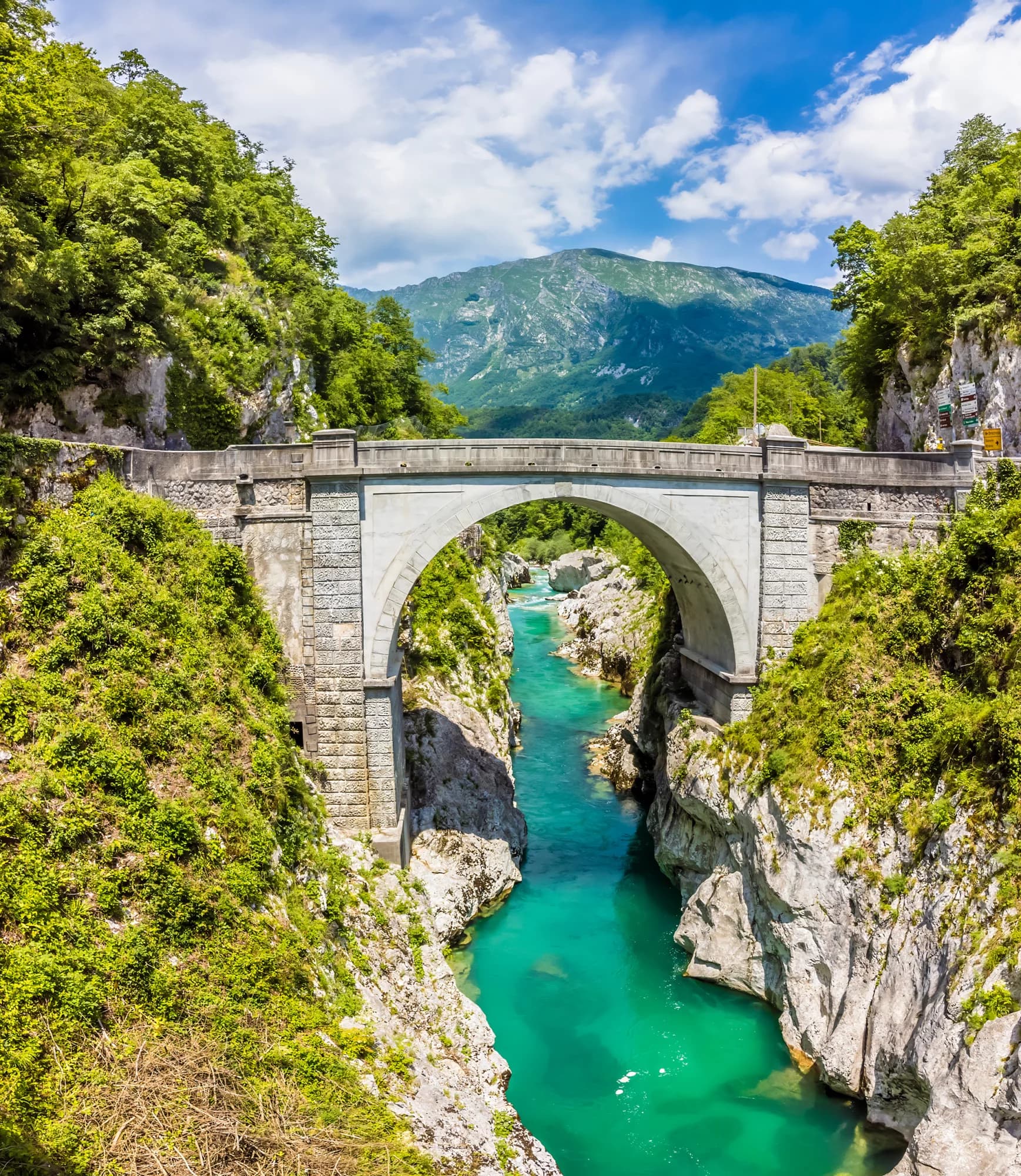 Napoleon Bridge over turquoise Soca River gorge with lush green mountains under a blue sky.