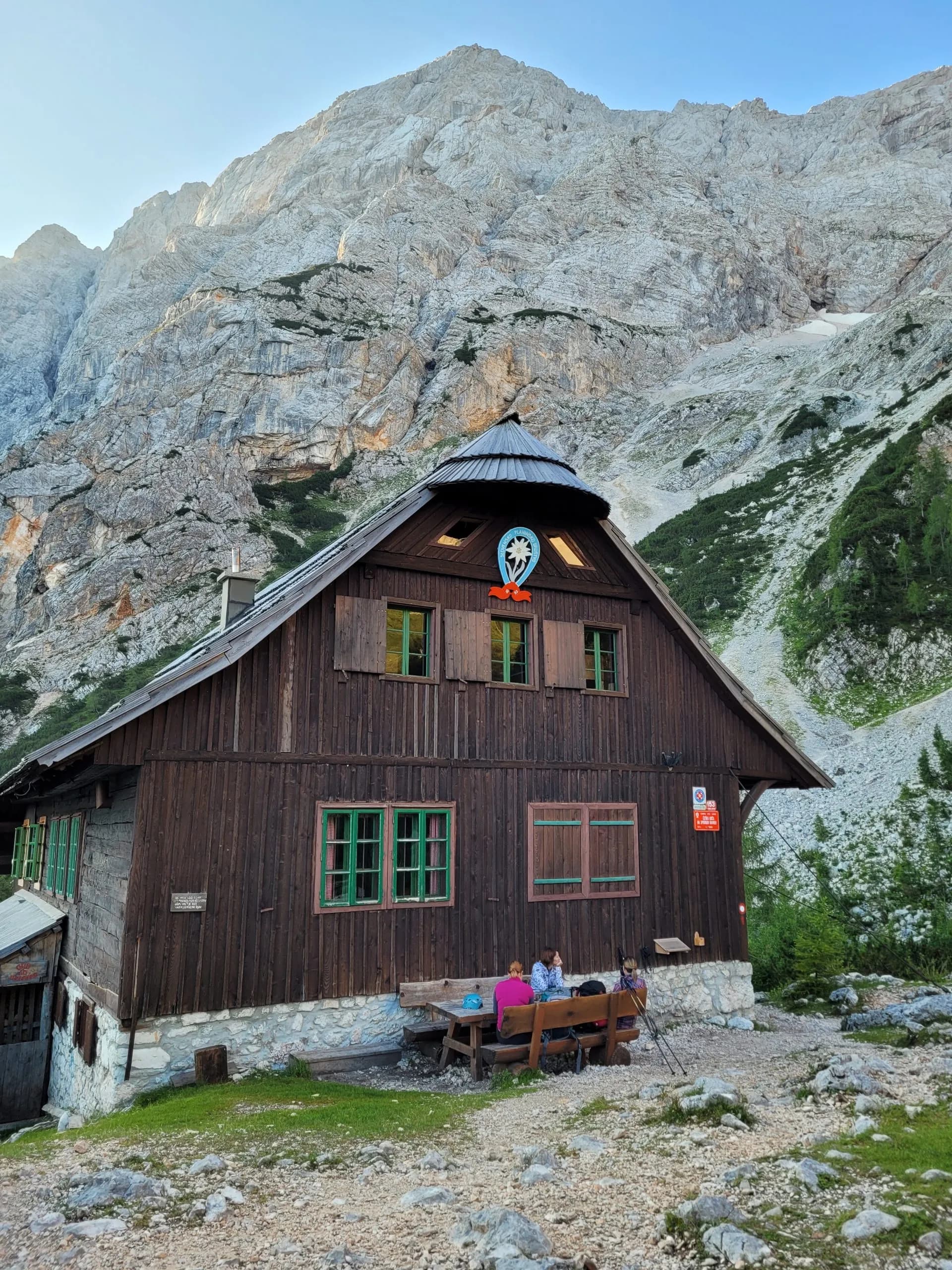 Mountain hut with hikers resting outside below massive, light-colored rocky mountains.