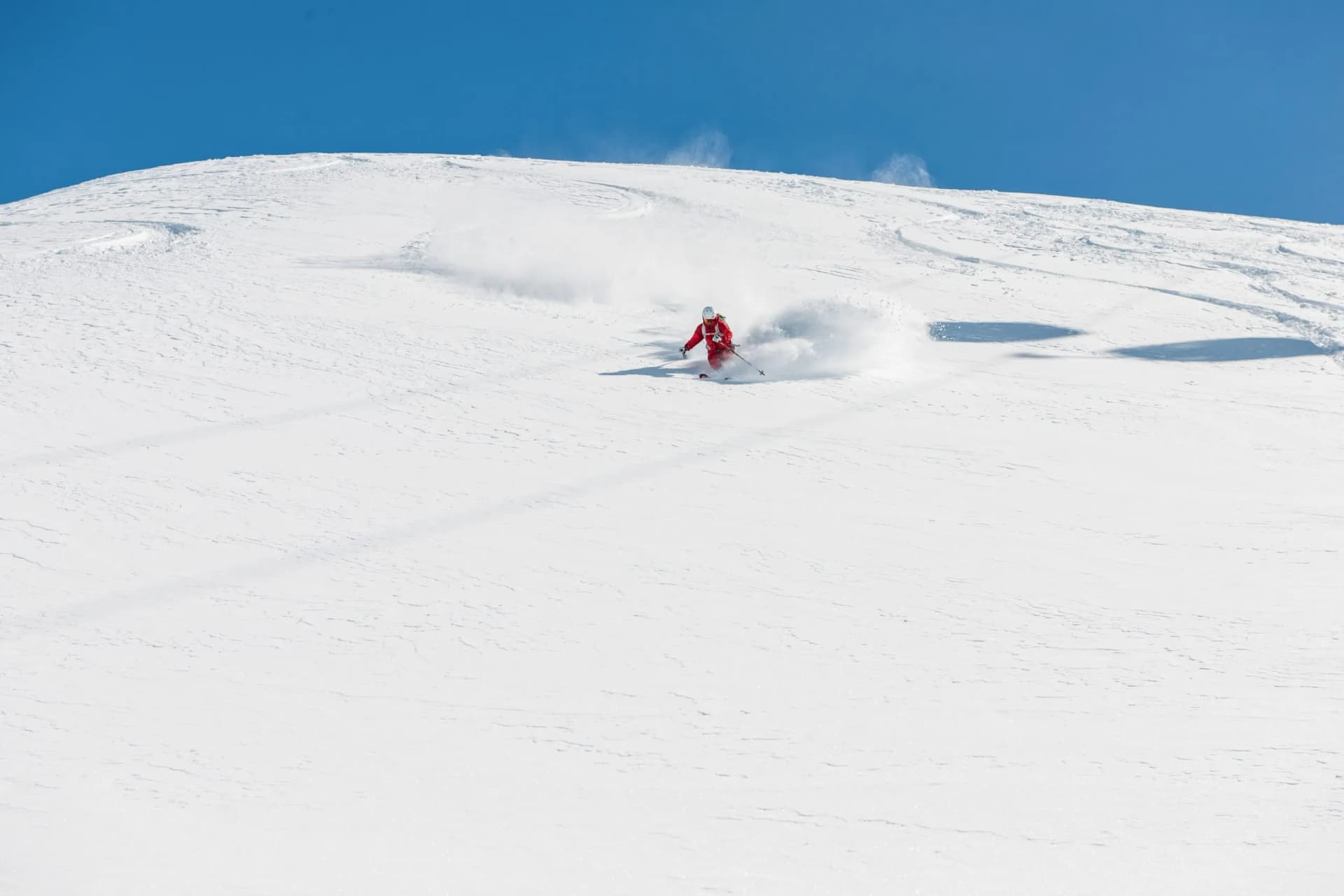 Skier in red suit making powder spray down a vast, snow-covered slope under a clear blue sky.