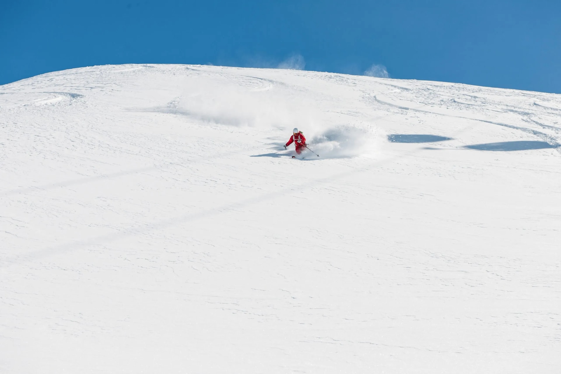Skier in red suit making powder spray down a vast, snow-covered slope under a clear blue sky.