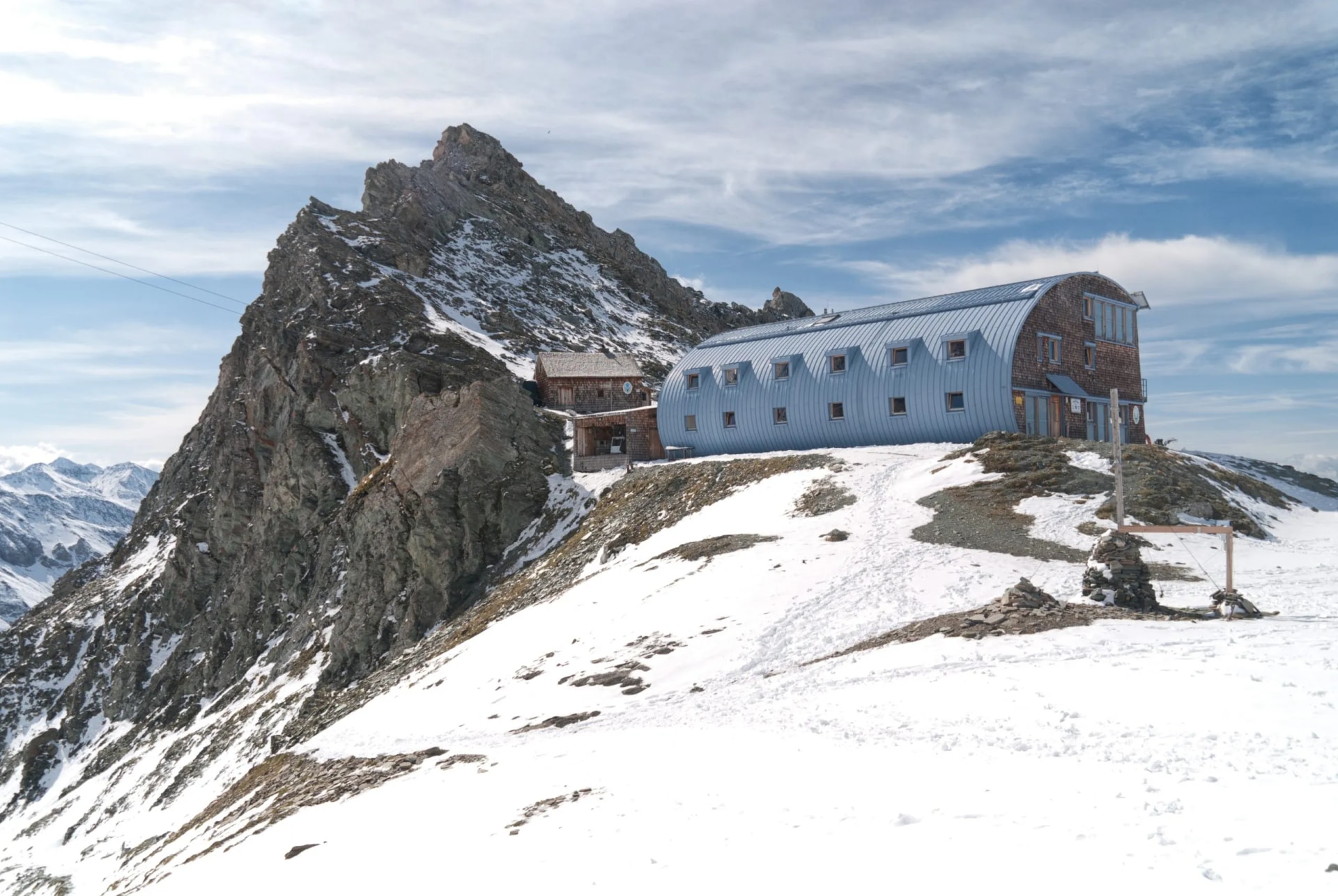 Alpine mountain hut with blue curved roof next to rocky peak in snow.