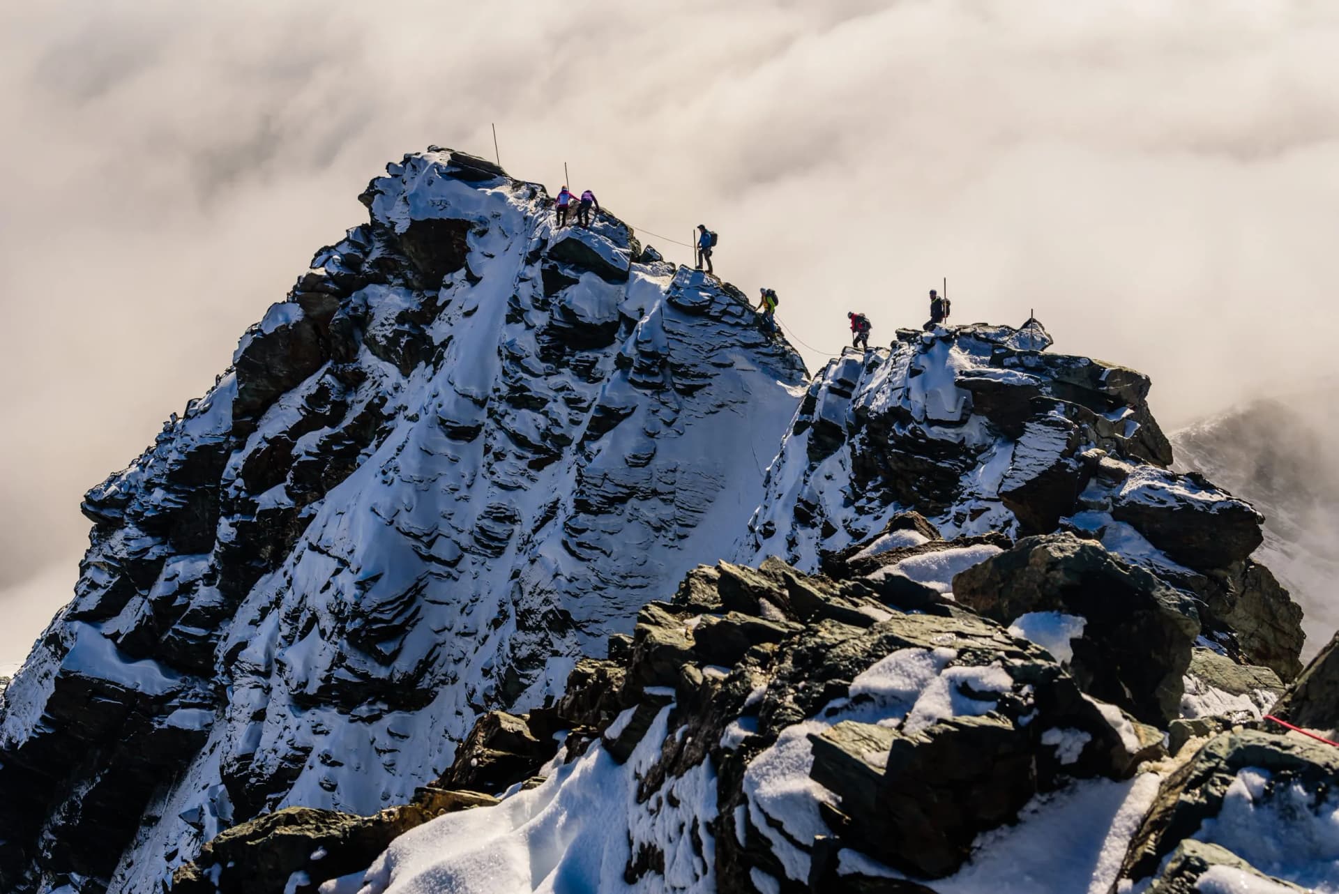 Mountaineers climbing a snowy, rocky ridge on the Grossglockner peak above the clouds.