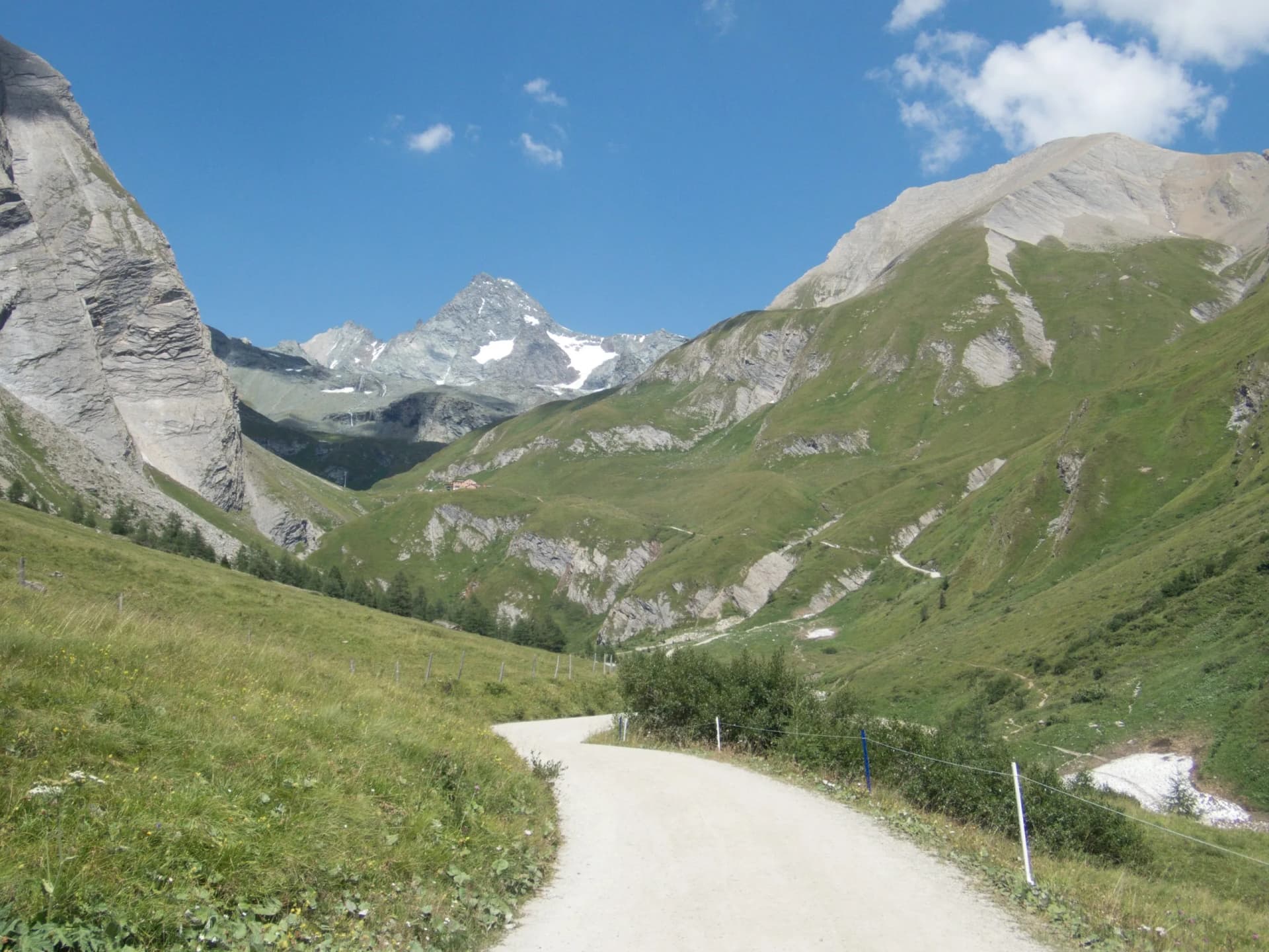 Unpaved road winding through green alpine valley toward snow-capped mountains under blue sky.
