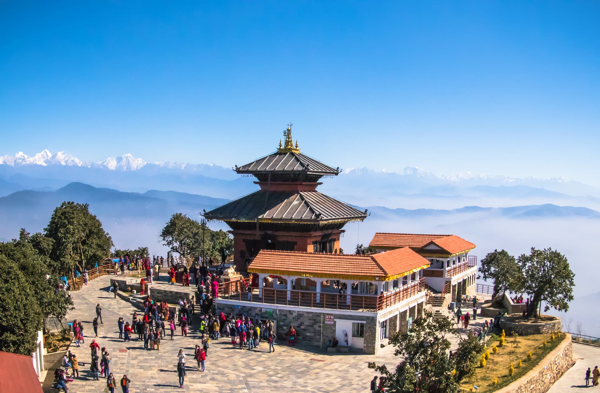 Bhaleshwor Mahadev temple complex with crowds, overlooking snowy Himalayan peaks.