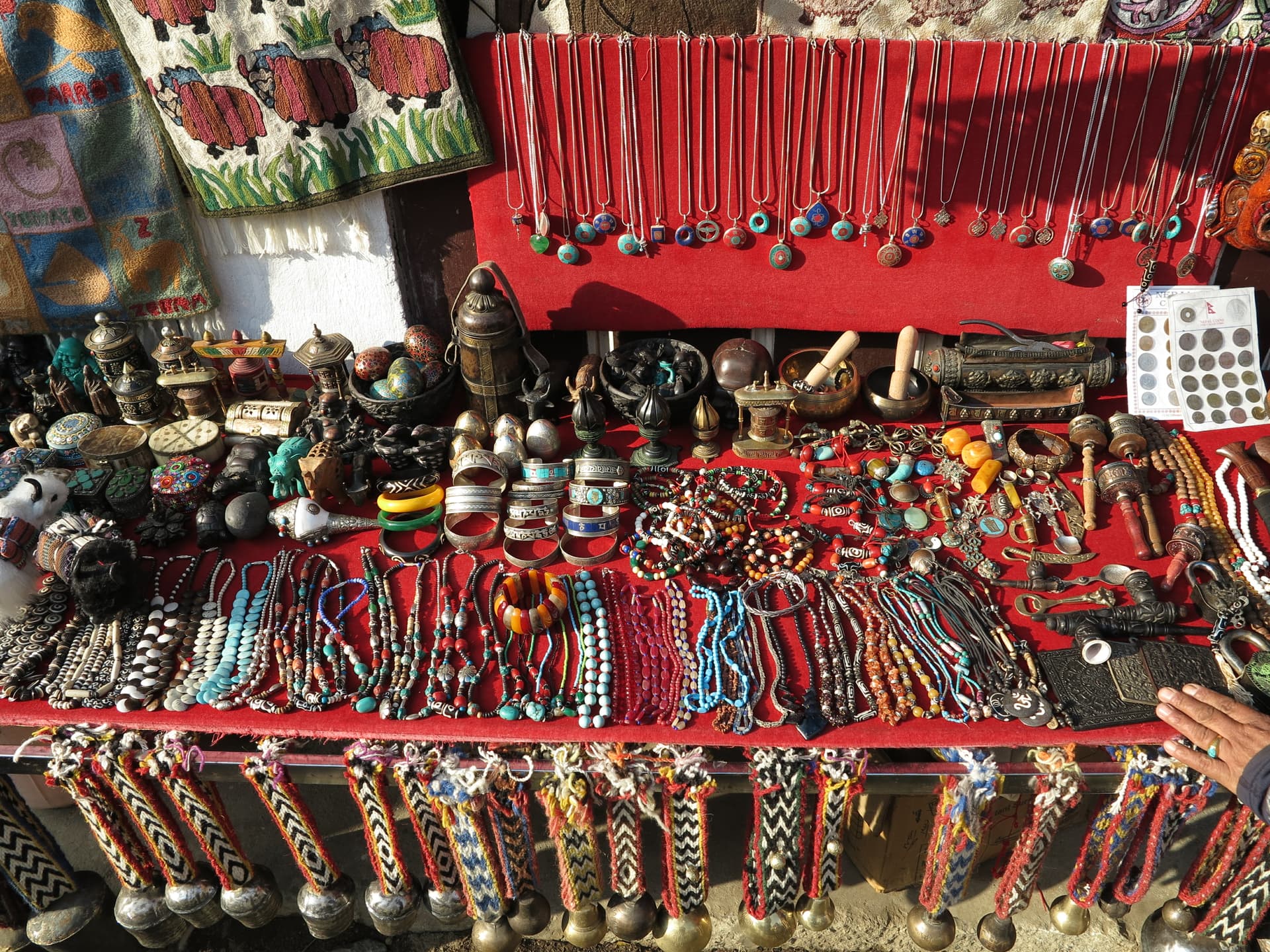 Jewelry and crafts stall with necklaces, bracelets, bells, and metal objects displayed on red cloth in Namche Bazaar.