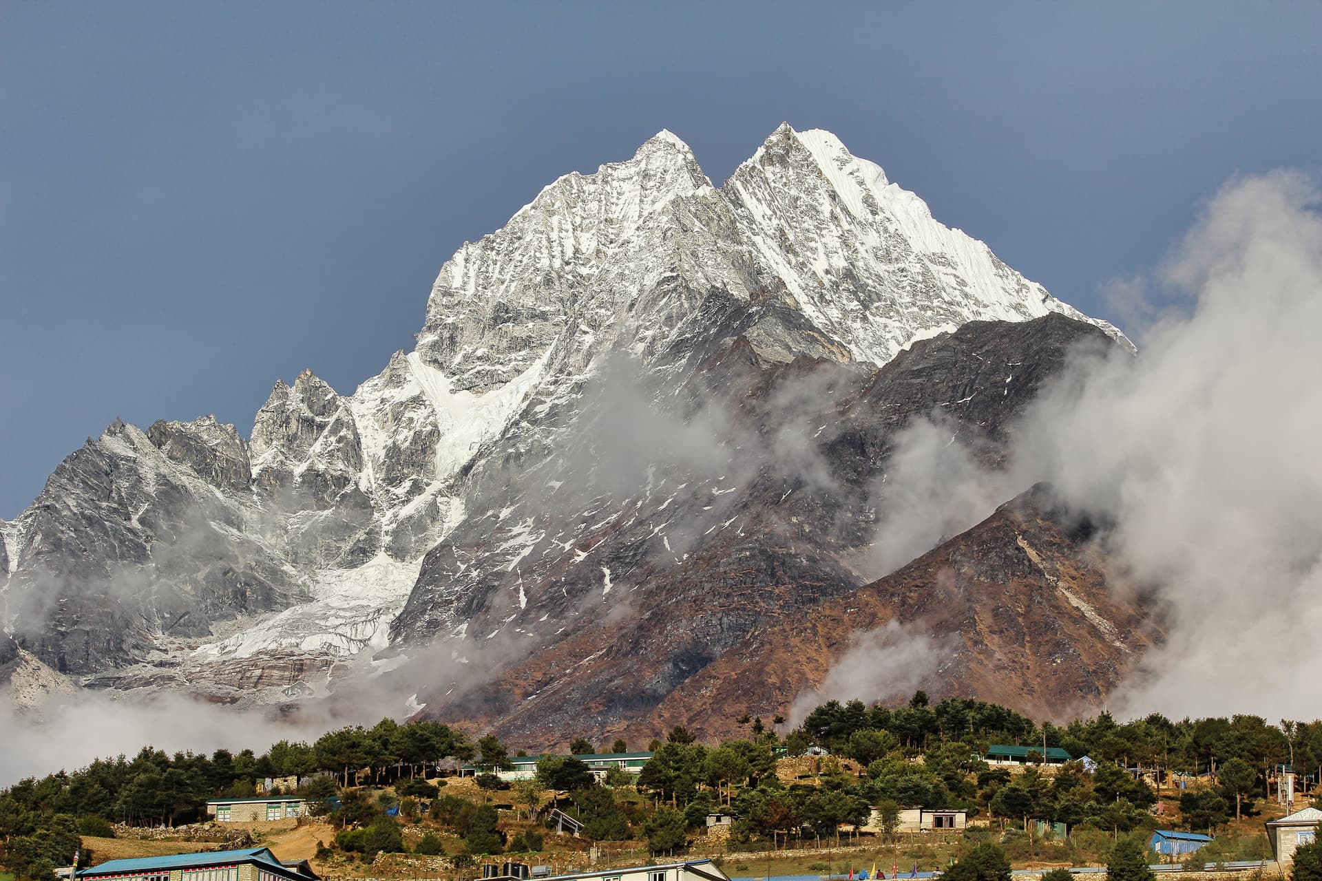 Snow-capped Thamserku twin peaks above village buildings shrouded in clouds in the mountains.