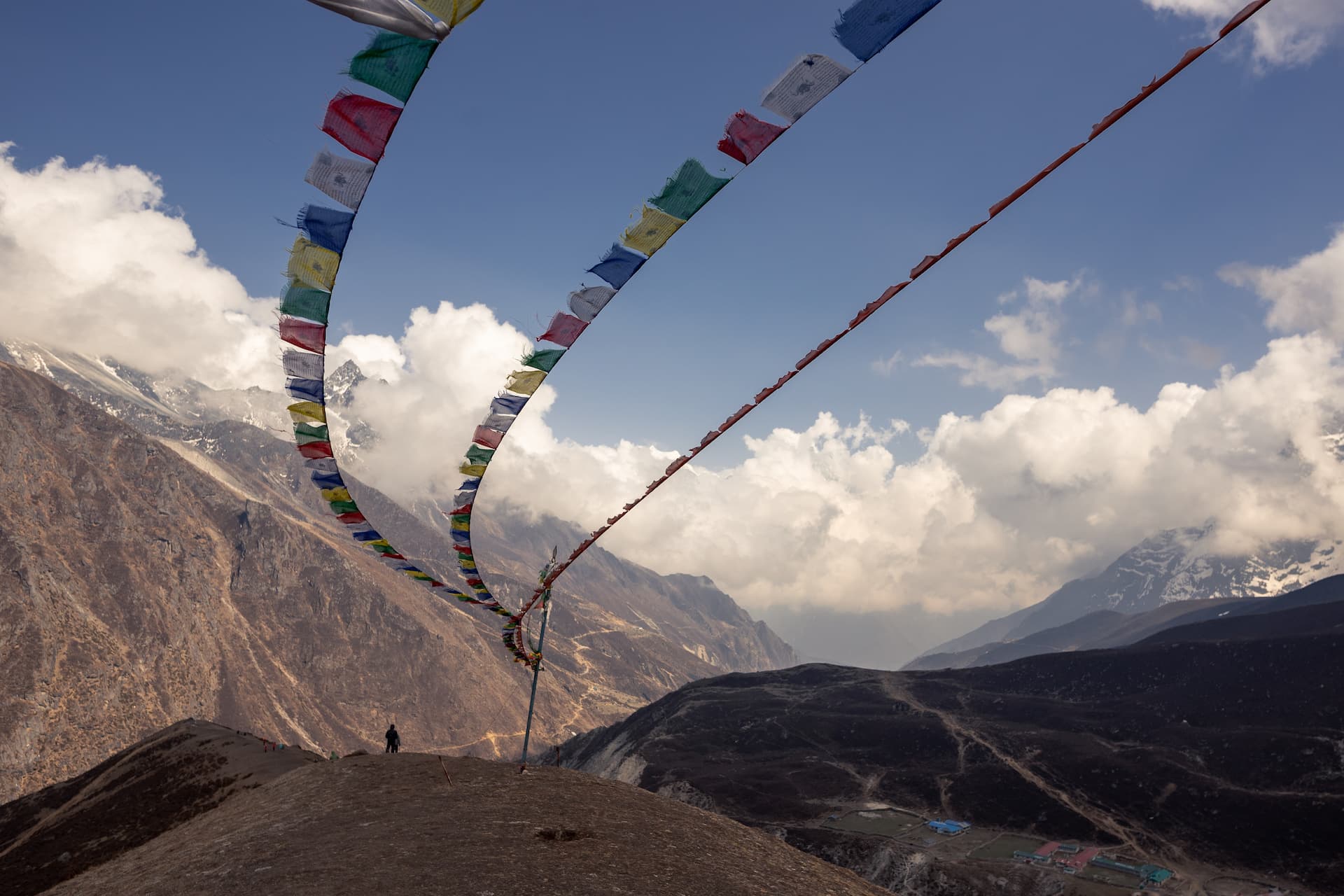 Prayer flags strung across mountain pass with hiker overlooking valley and snow-capped peaks.
