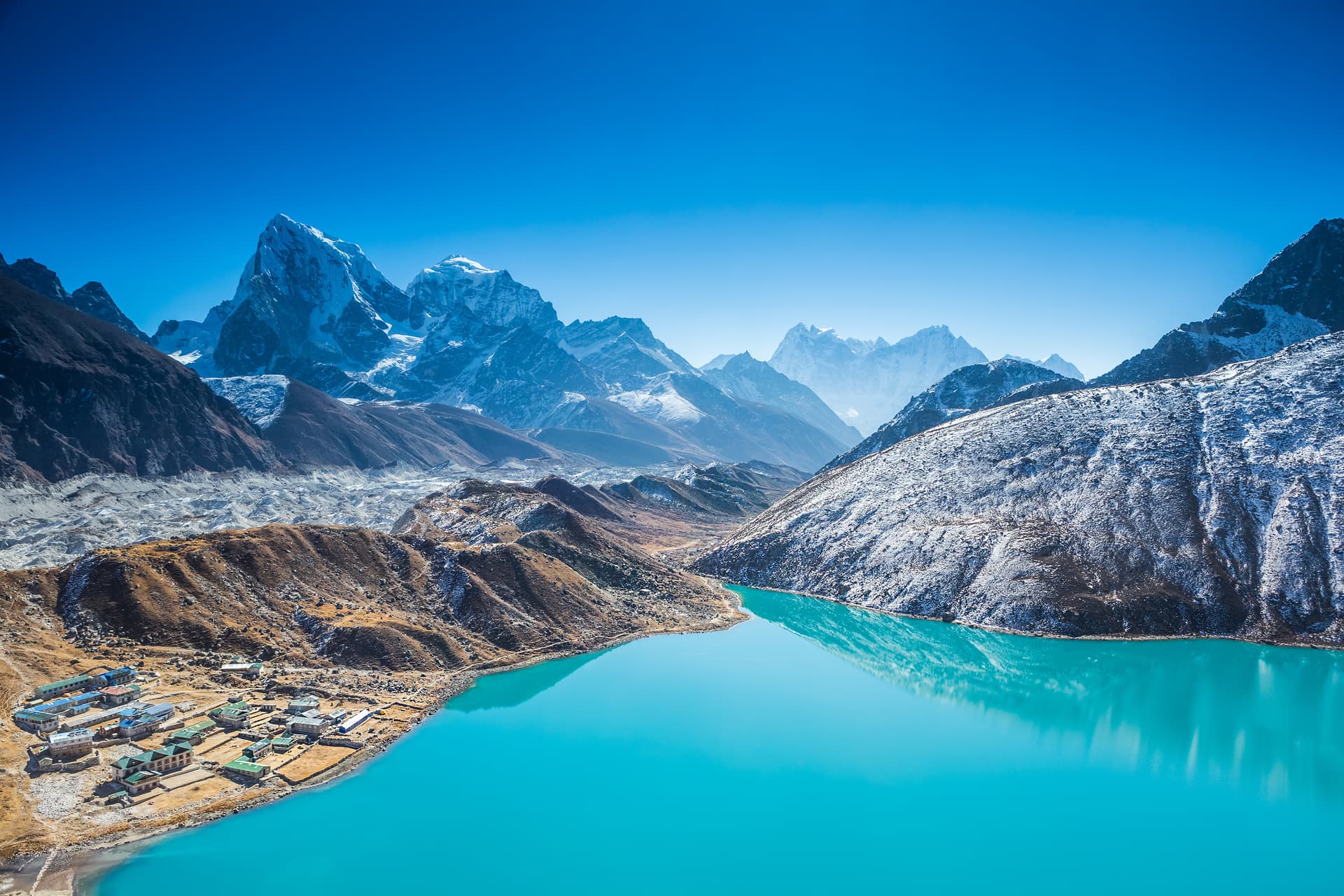 View from Gokyo Ri showing turquoise Gokyo Lake, village, glacier, and snow-capped Himalayan mountains.