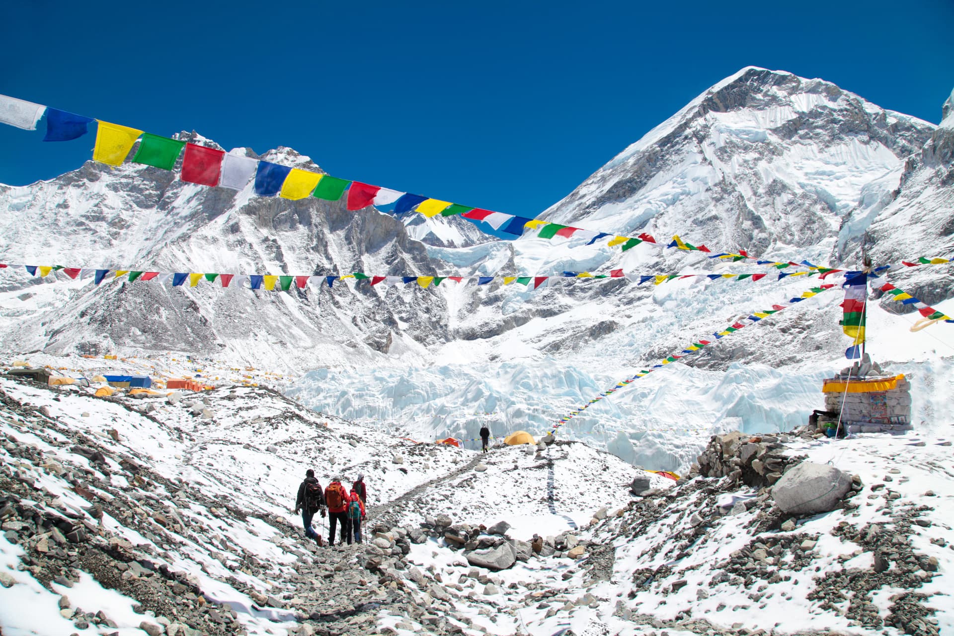 Hikers trekking past prayer flags at Everest Base Camp with snow-covered mountains and glacier.