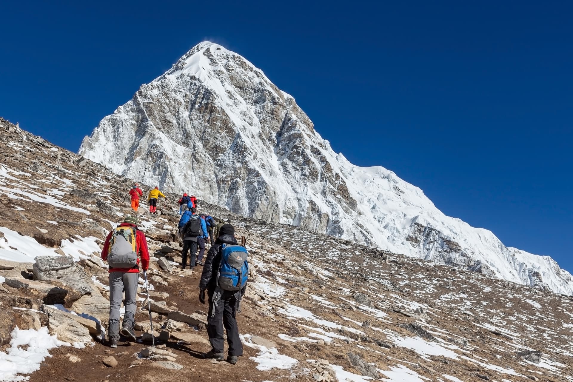 Group of trekkers hiking up a rocky slope toward a massive snow-covered mountain under a clear blue sky.