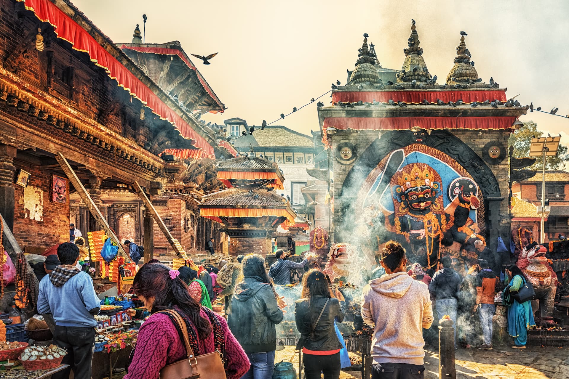 Crowd gathered at Kathmandu center market near temple gate with smoke and colorful deity carving.