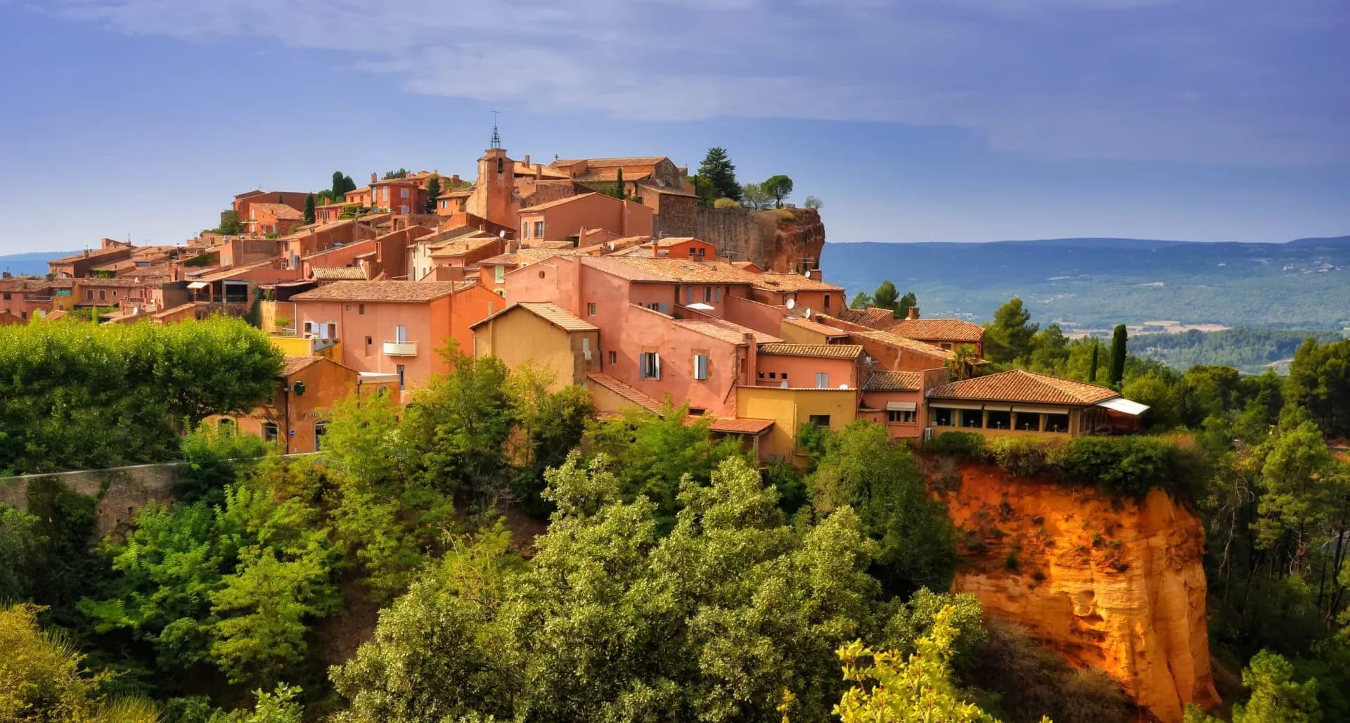 Village with ochre-colored houses perched on a cliff overlooking a forested valley in Roussillon.