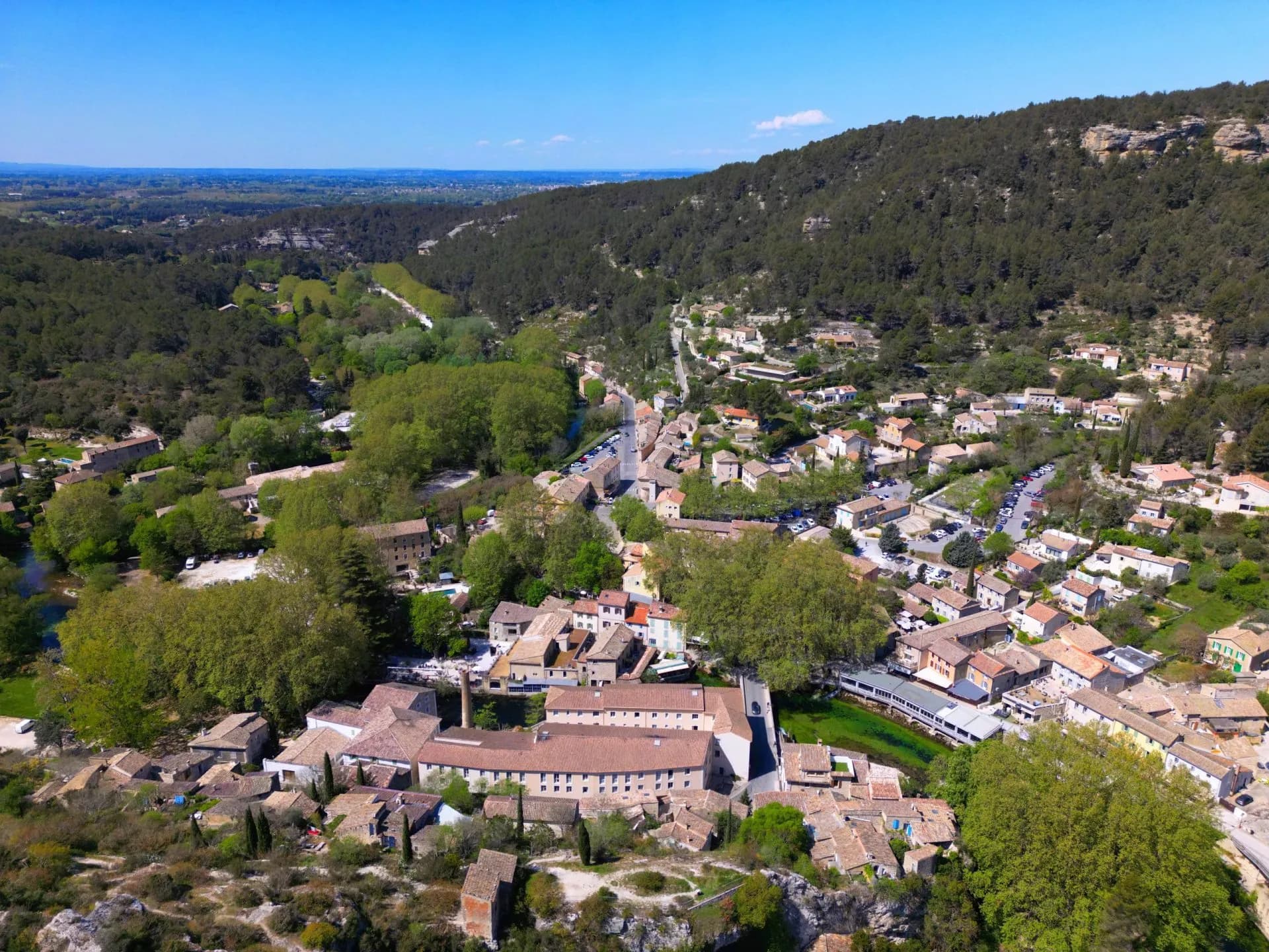 Aerial view of Fontaine-de-Vaucluse village nestled in a valley with dense forest and clear blue sky.